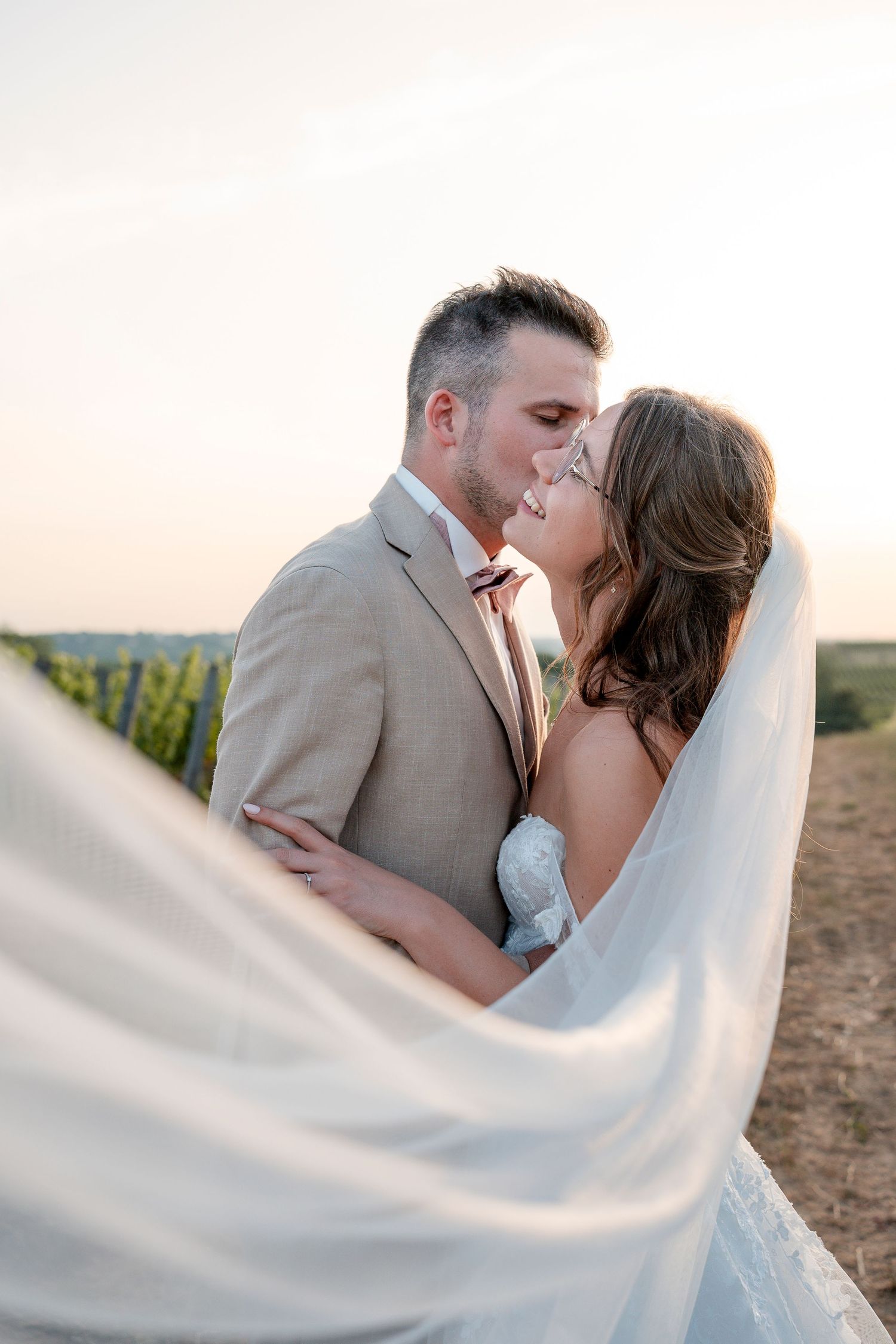 Bride and groom sharing a romantic moment outdoors at sunset, with veil flowing in the breeze near a vineyard.
