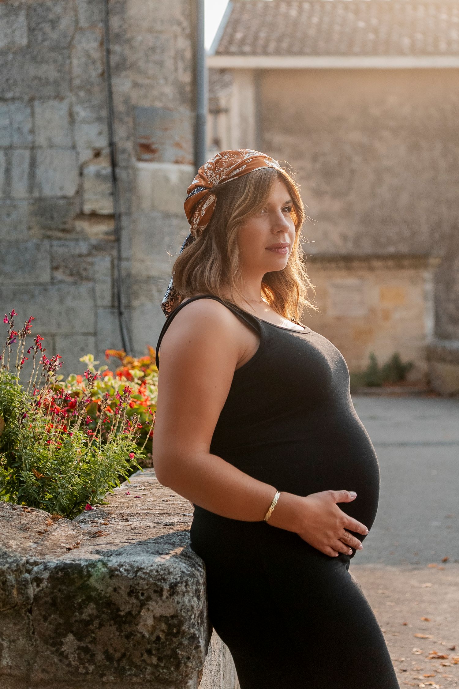 Maternity photos of an expectant mother in a black dress posing against a stone wall with colorful flowers.