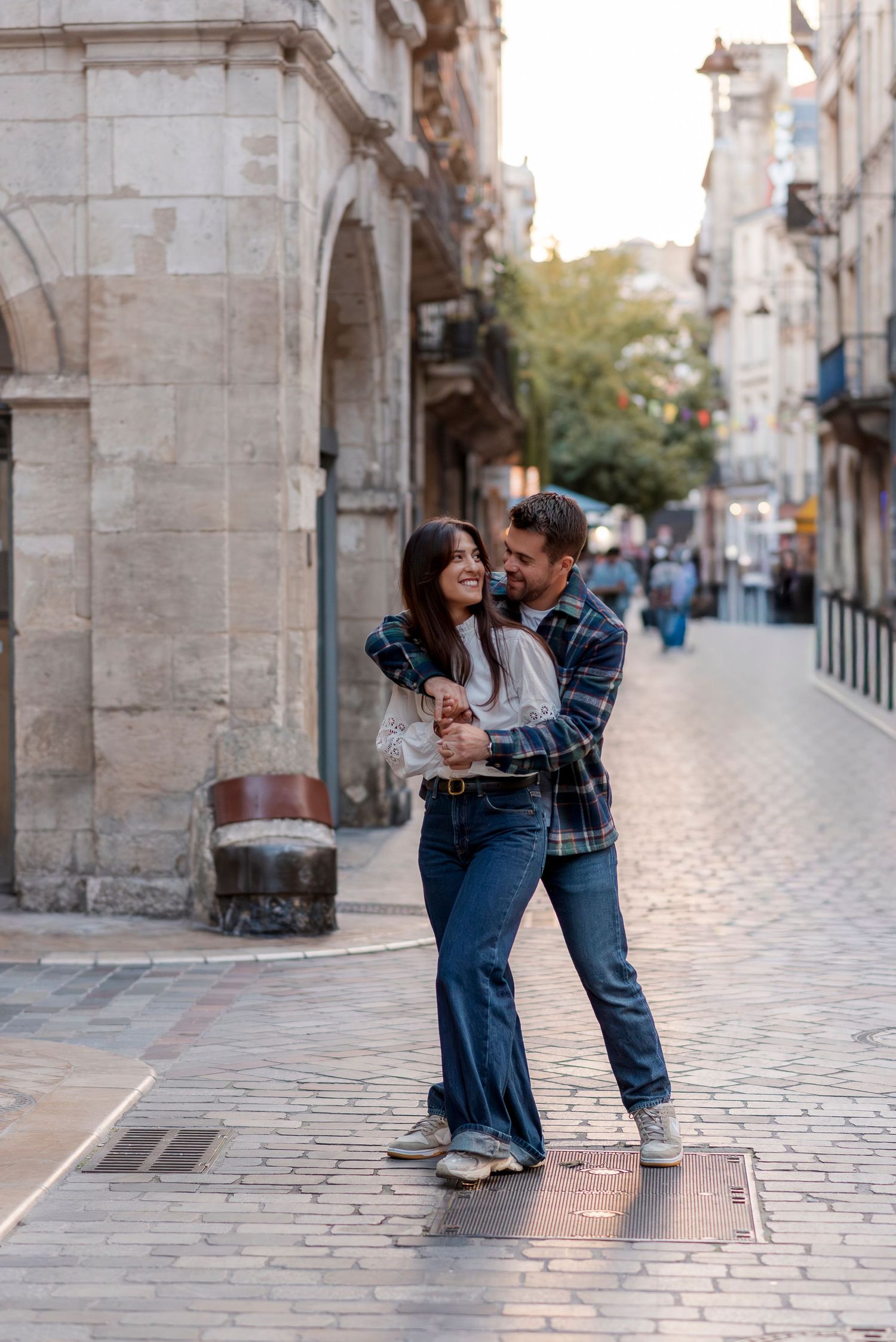 Happy couple embracing and dancing on a cobblestone street in a European city, both wearing jeans and casual tops.