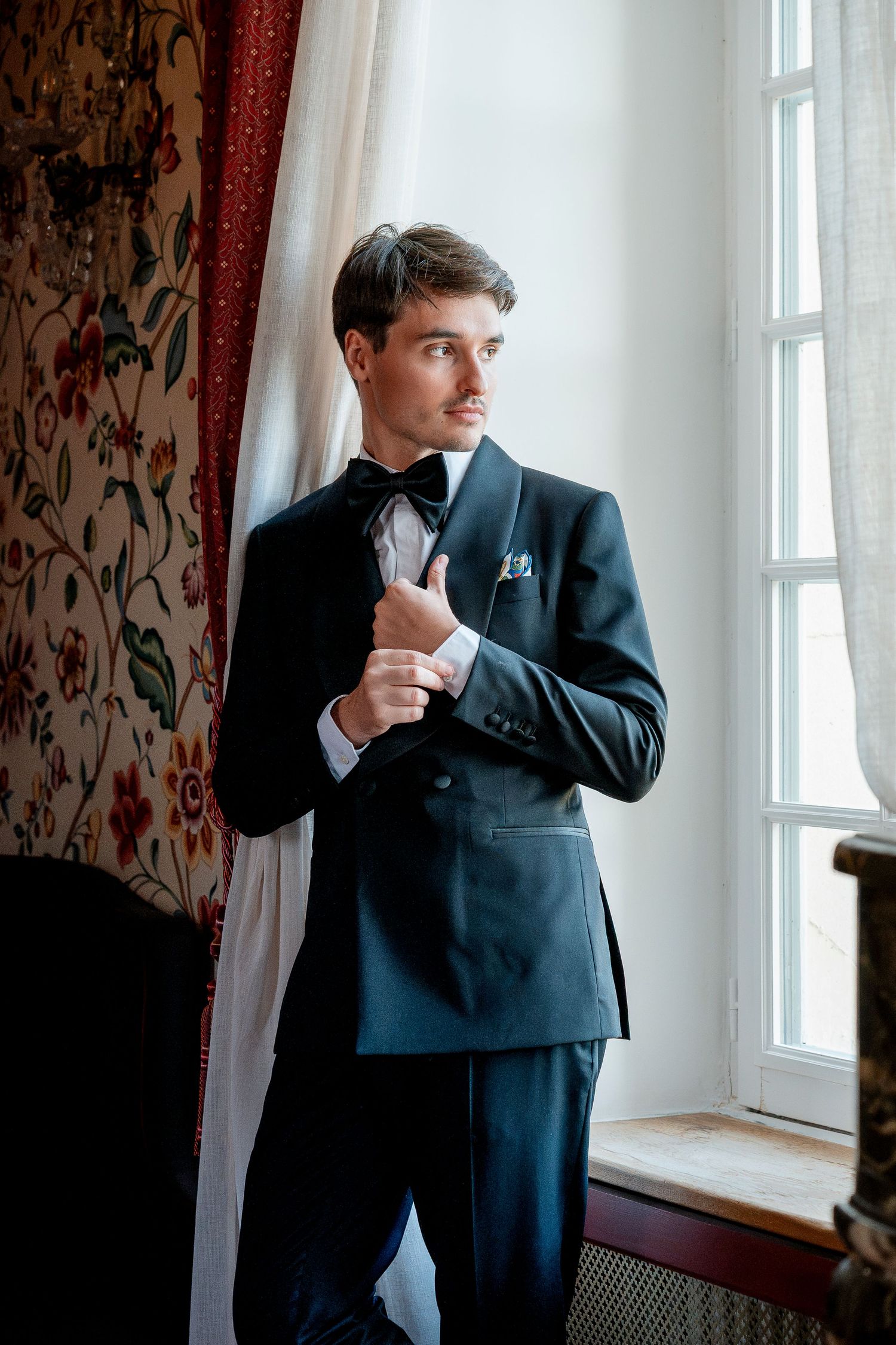 Man in a black tuxedo and bow tie adjusts his cuff while gazing out a sunlit window beside floral wallpaper and red curtains.