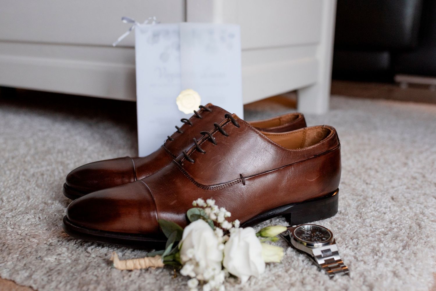 Brown leather dress shoes with white floral boutonniere arranged on carpet.