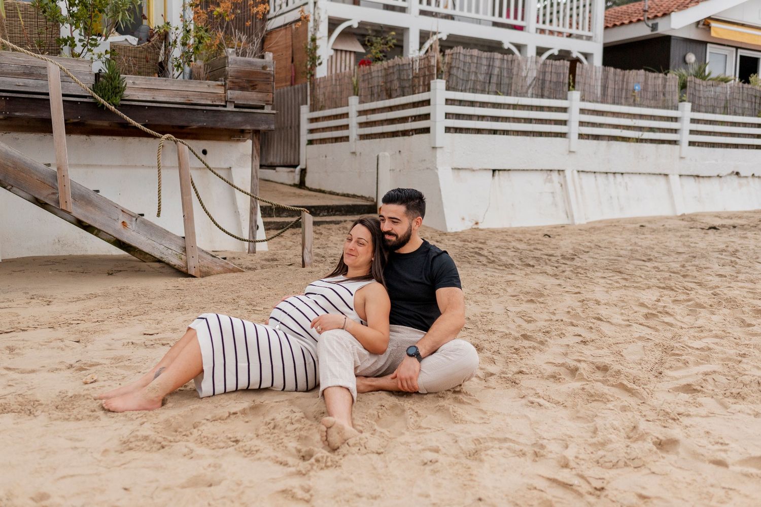 Couple lounging together on sandy beach with beach houses in background.