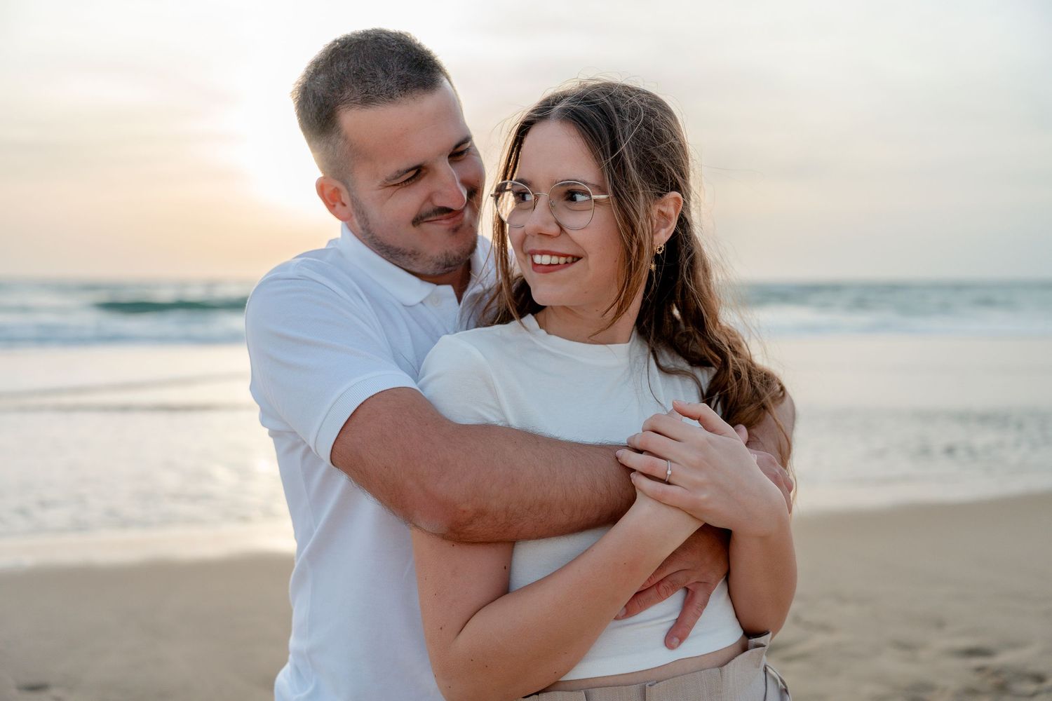 Couple embracing on beach at sunset, woman in glasses smiling, man hugging her from behind near ocean waves.