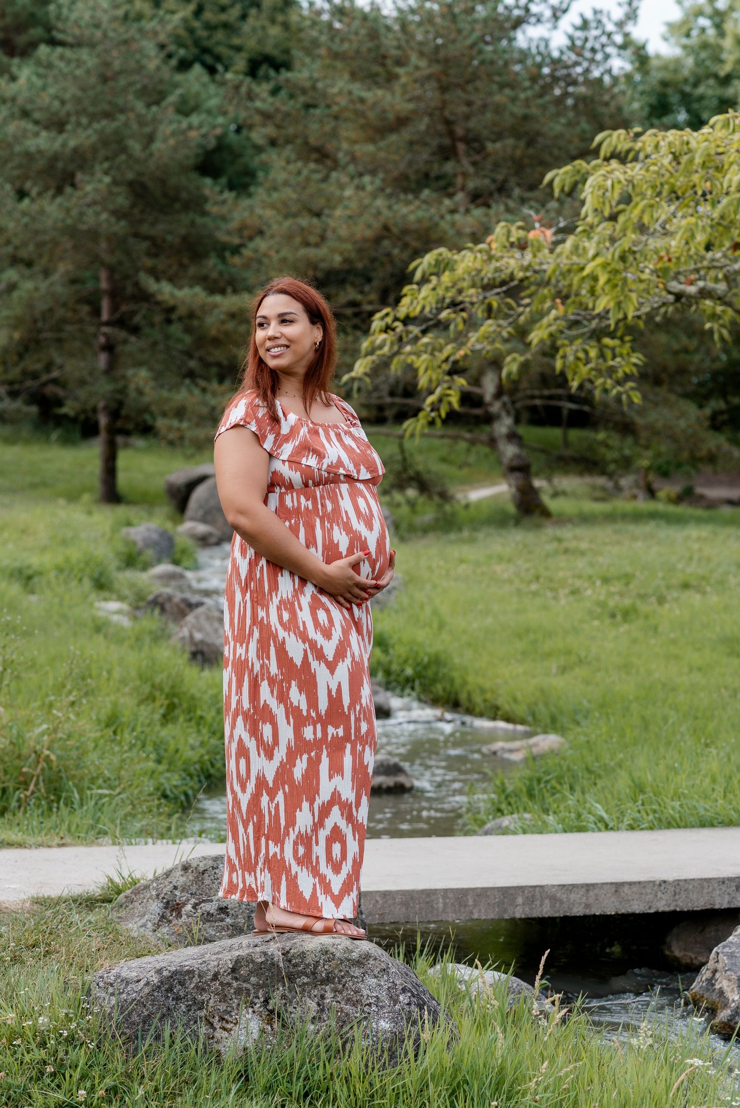 A person in an orange and white patterned dress poses by a stream in a lush green outdoor setting.