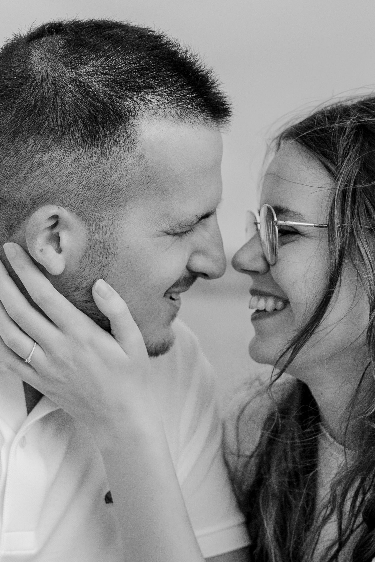 Black and white photo of a smiling couple facing each other closely, woman wearing glasses, man wearing a ring.