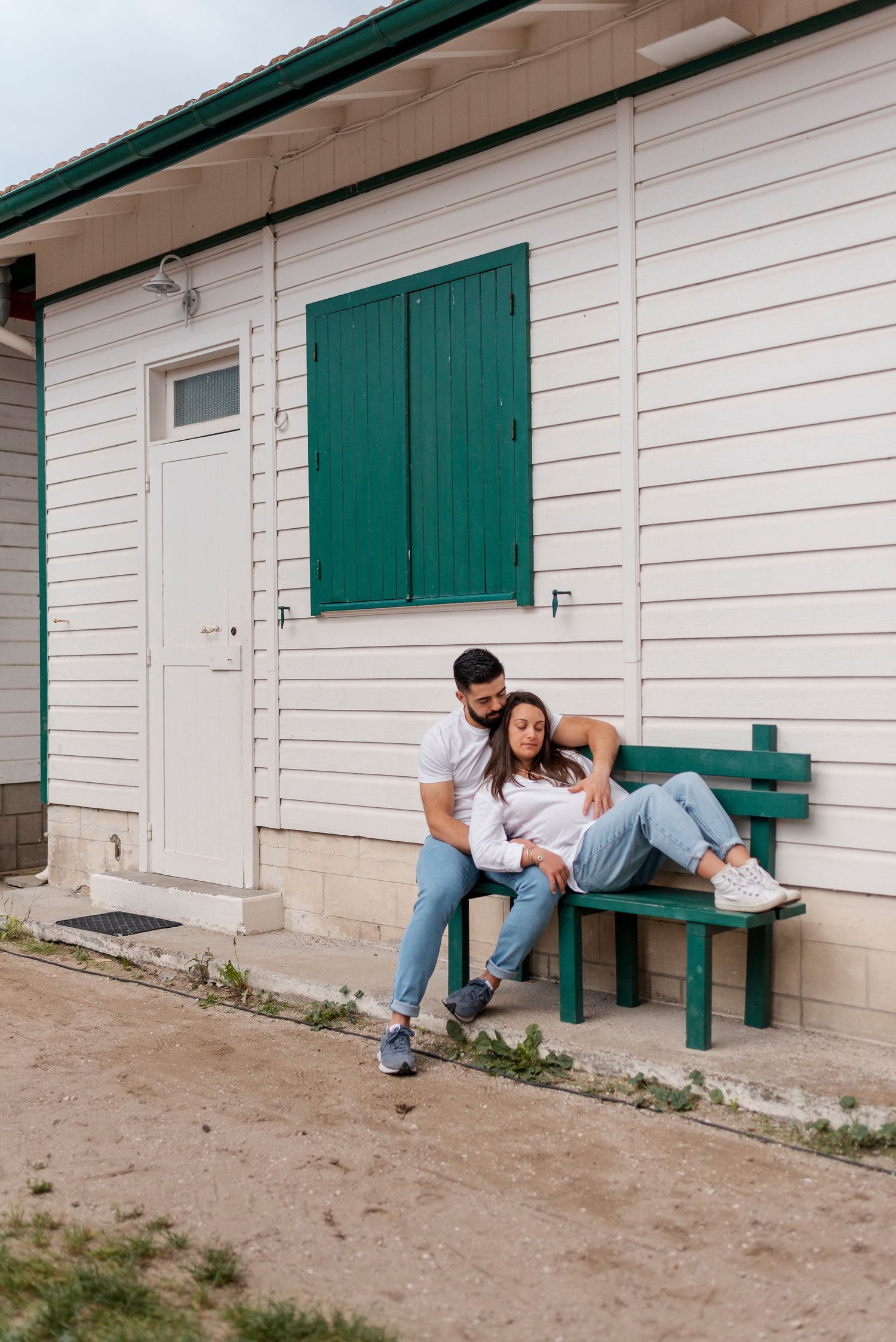 Couple relaxing on a green bench against white building with teal shutters.