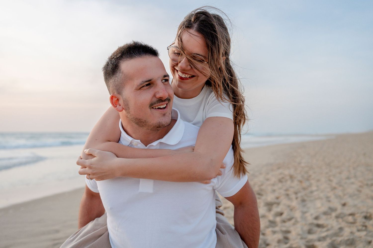 A happy couple enjoys a piggyback ride on a sunny beach, smiling and embracing near the ocean waves.