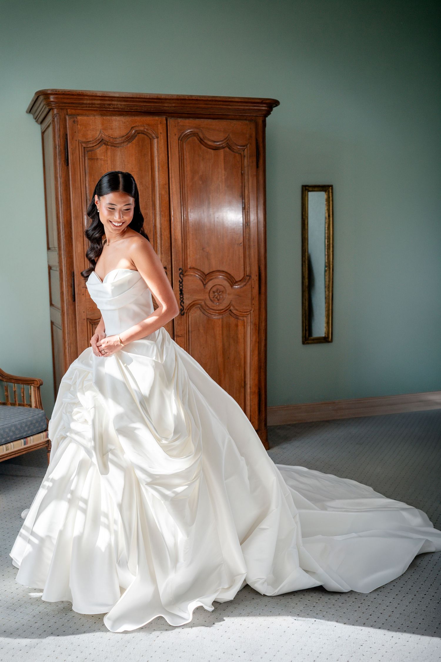 Smiling bride in a strapless ivory satin ball gown with a long train stands beside a carved wooden armoire in a sunlit room.