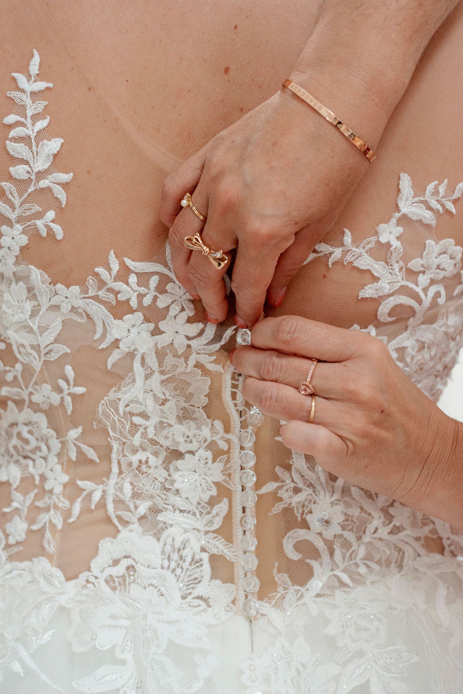 Close-up of delicate white lace detail on a wedding dress with hands showing rings.
