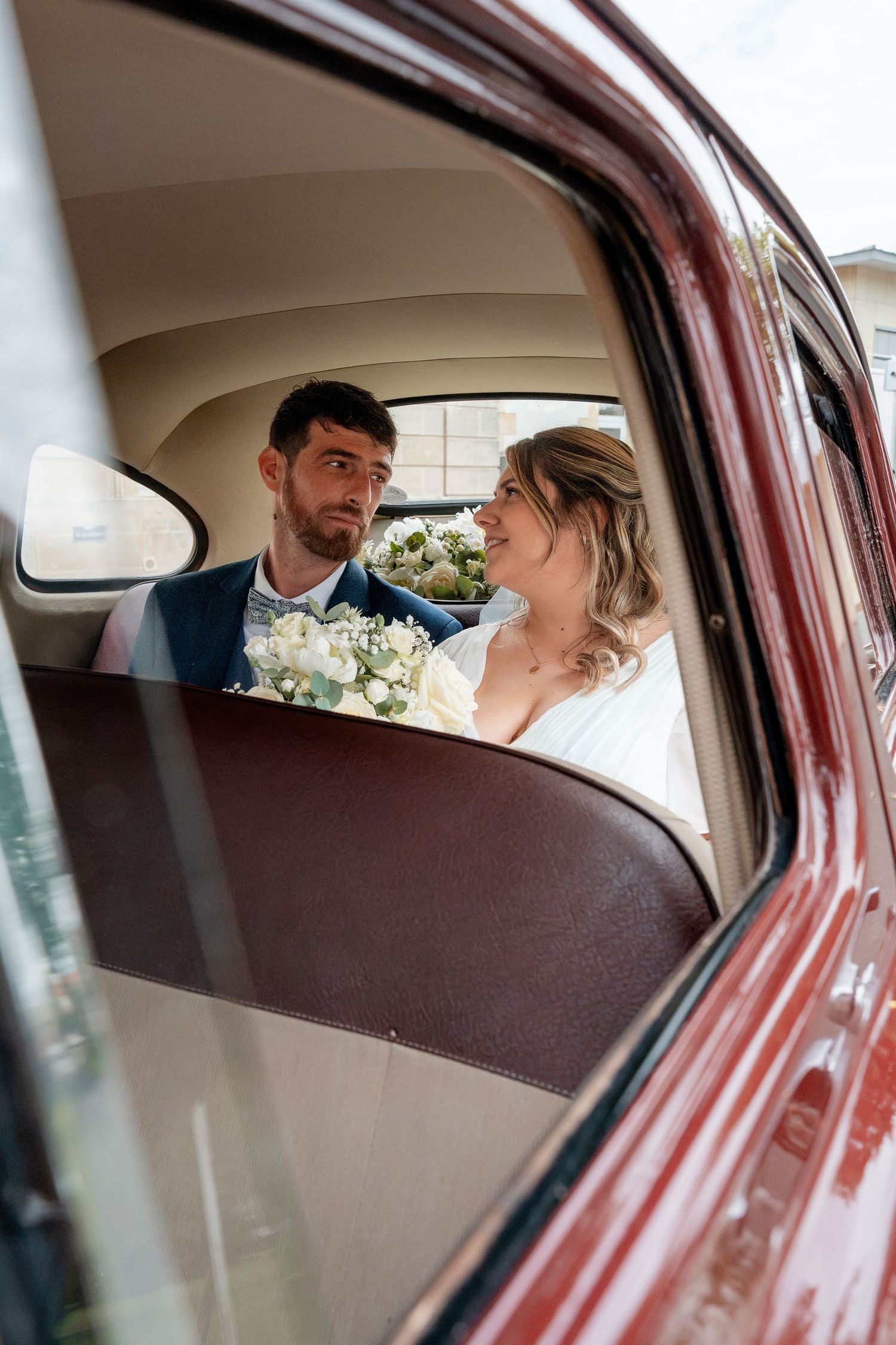 Smiling bride and groom sit together in the back seat of a vintage red car, holding a white floral wedding bouquet.