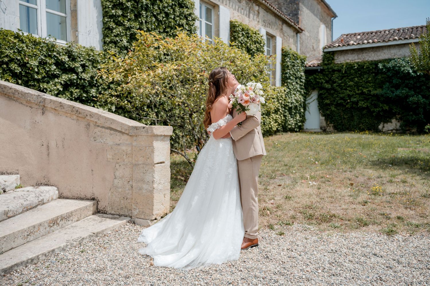 Bride in a flowing white gown and groom in a tan suit embrace outdoors beside stone steps, hedges and a historic building.
