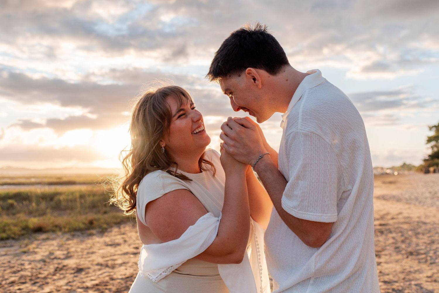 A couple shares a romantic moment on a sandy beach at sunset, both dressed in white casual attire.