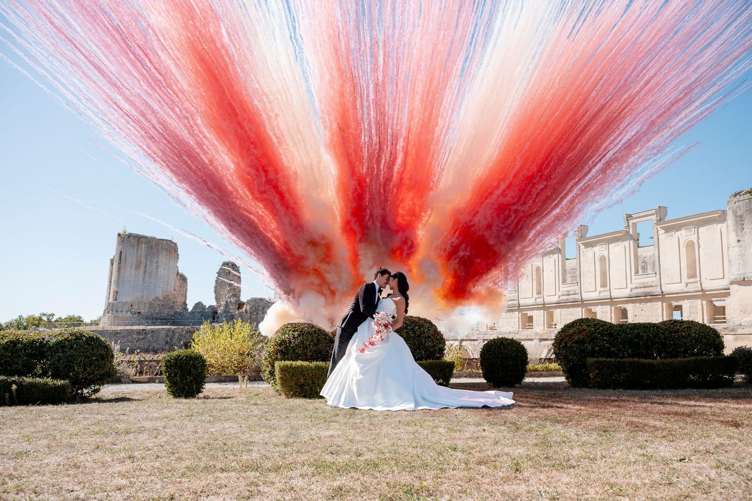 Bride in a white gown and groom in a dark suit embrace in a garden as red and white fireworks burst above historic stone buildings.