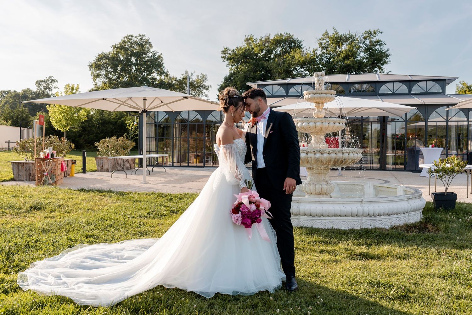 Newlyweds share intimate moments by ornate stone fountain in garden setting.