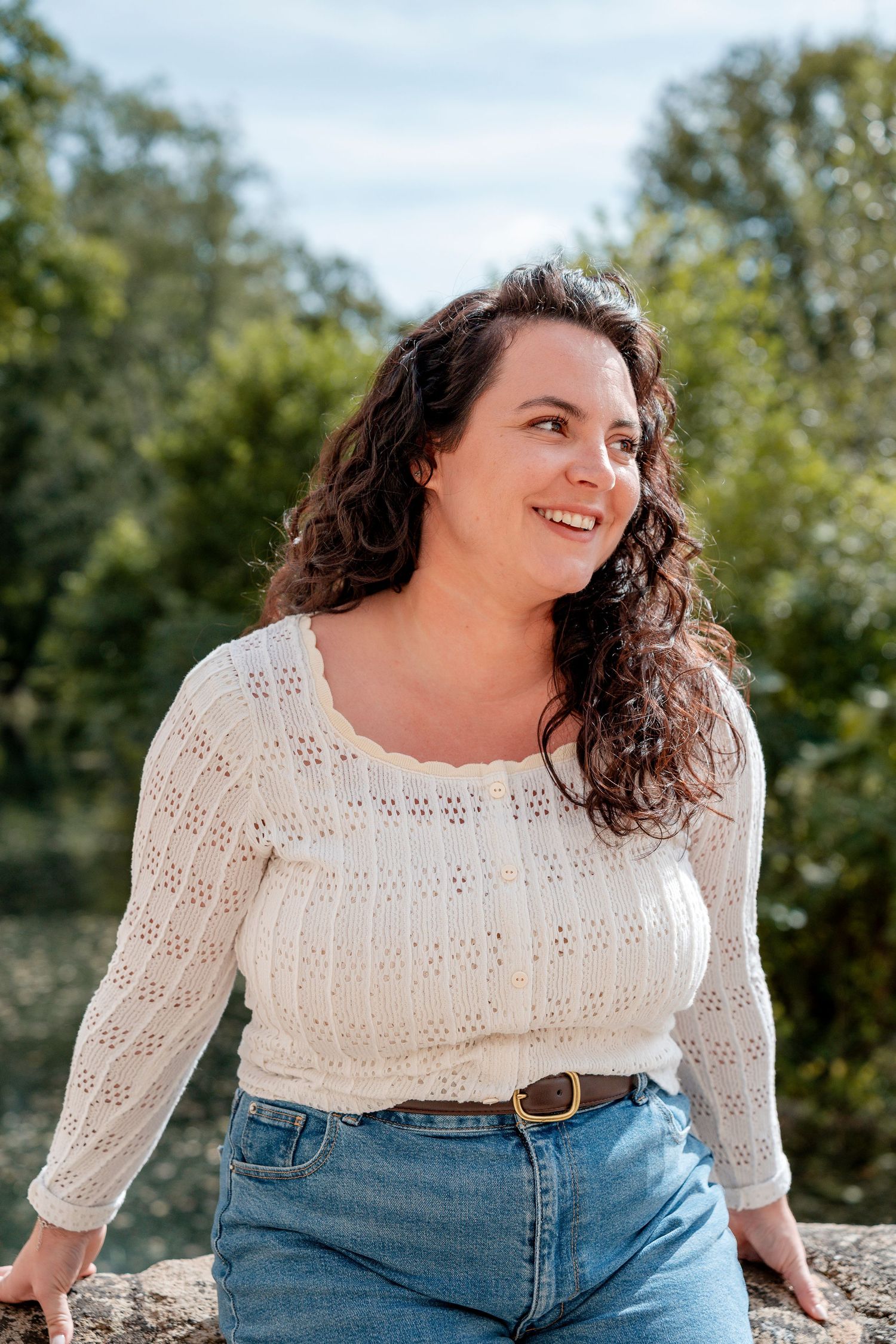 Smiling woman with curly dark hair wearing a white lace top and jeans, posing outdoors with green trees behind her.