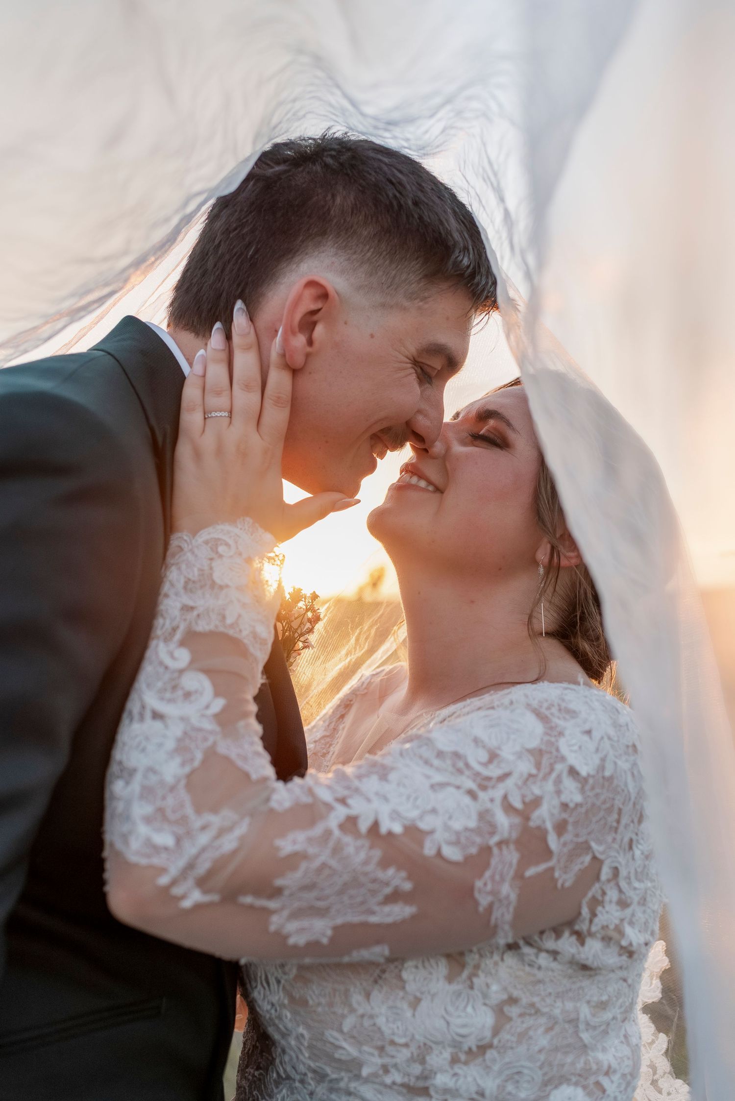 Romantic wedding couple sharing intimate kisses under a veil with golden sunlight glowing behind them.