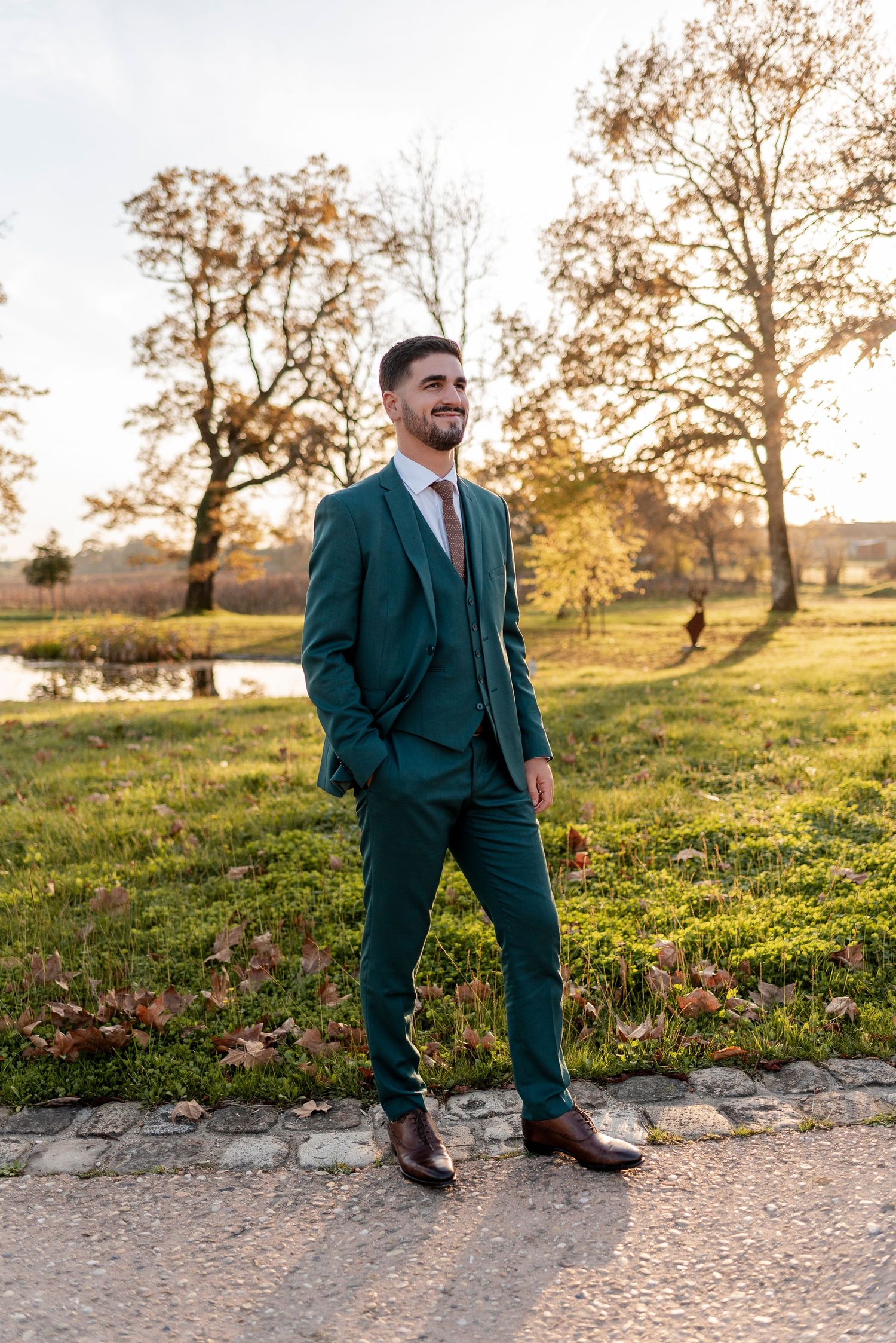Groom in dark green suit poses on stone wall during sunset outdoor photo session.
