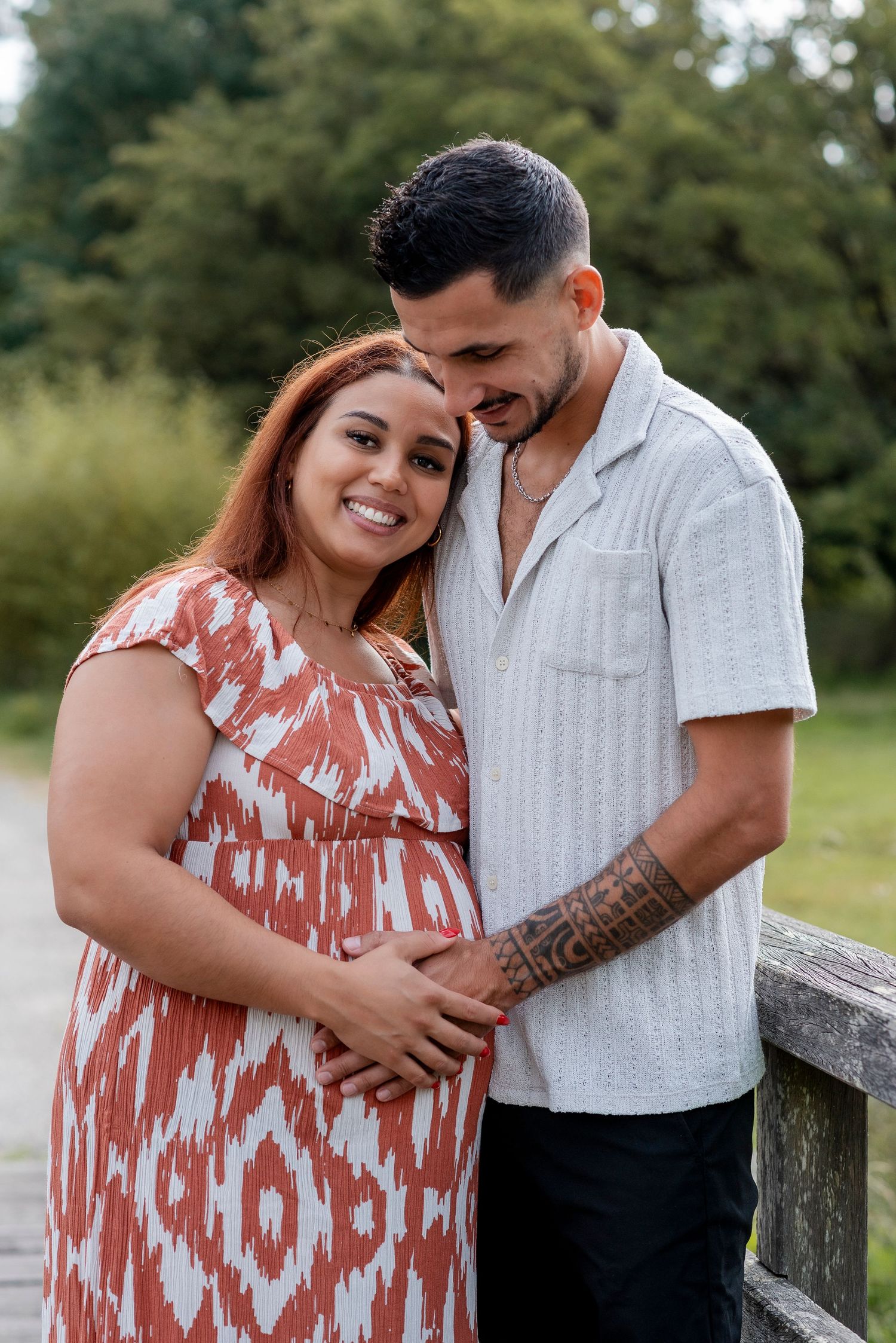 A couple embraces while standing on a wooden bridge over a stream in a natural setting.