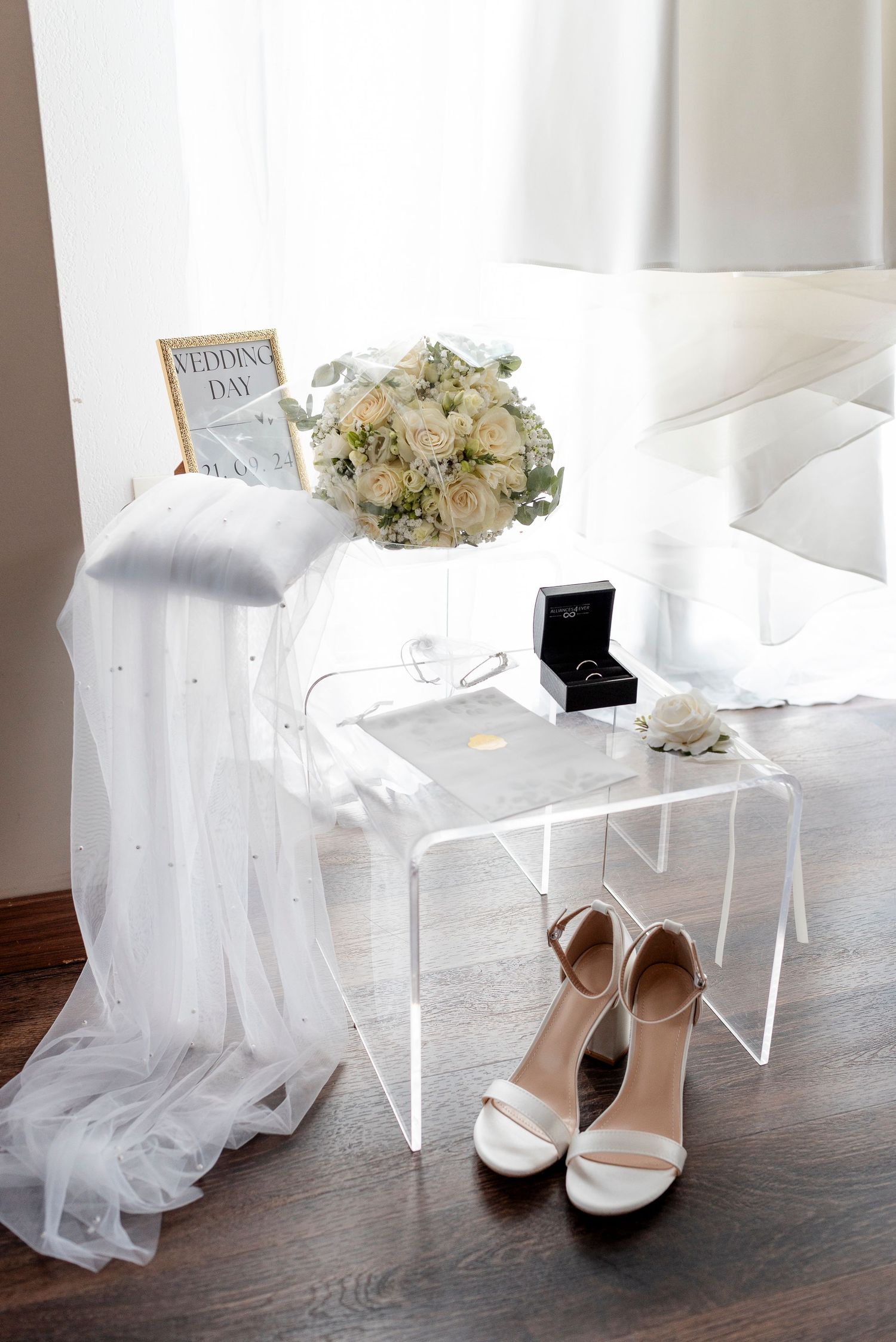 Elegant white wedding shoes and veil arranged on a dark wooden floor with delicate floral bouquet.