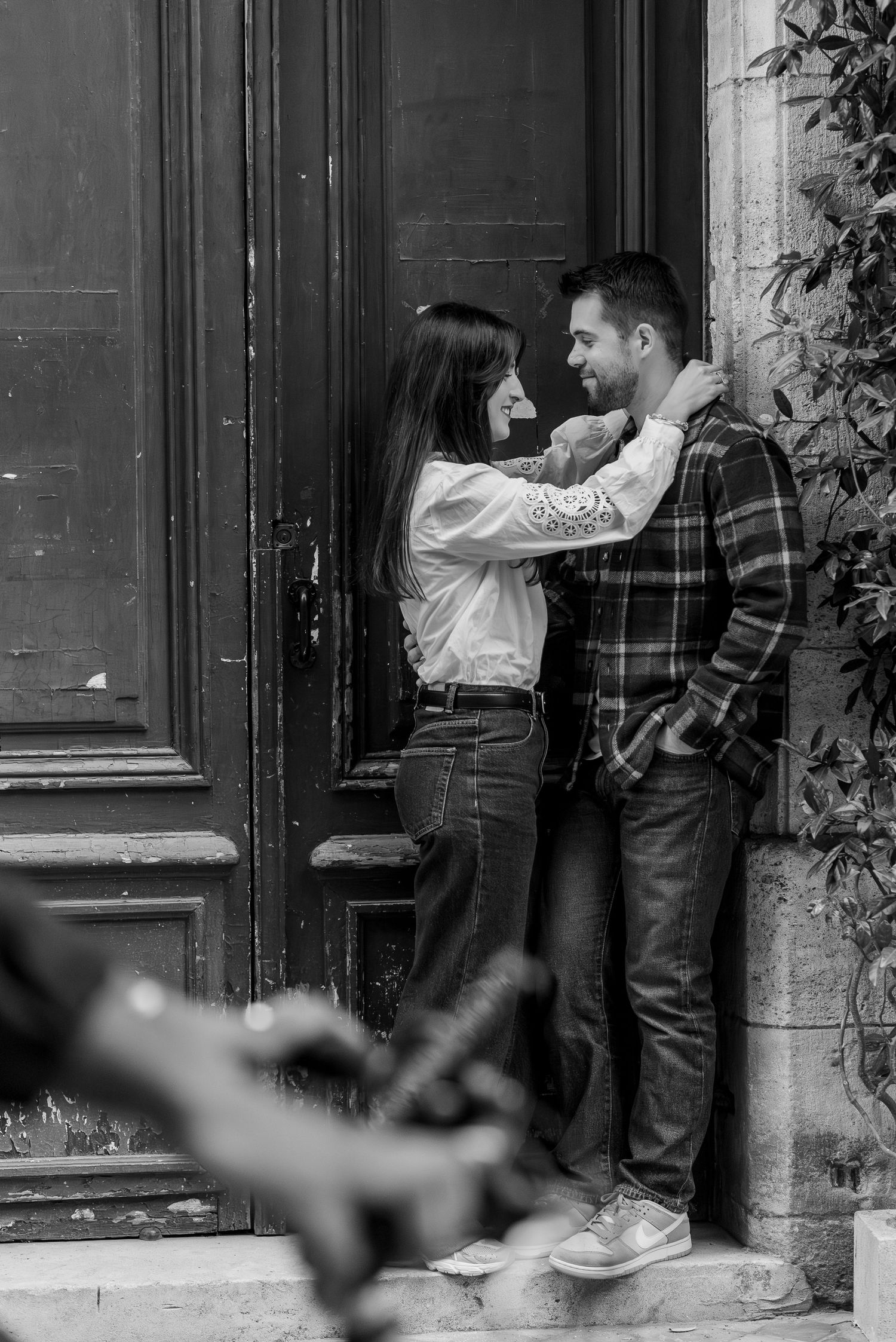 A couple poses playfully against a dark wooden door in black and white.