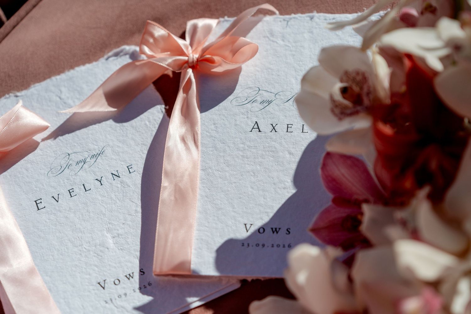 Handmade paper wedding vow books for Evelyne and Axel tied with peach satin ribbon beside pink and white orchid flowers.