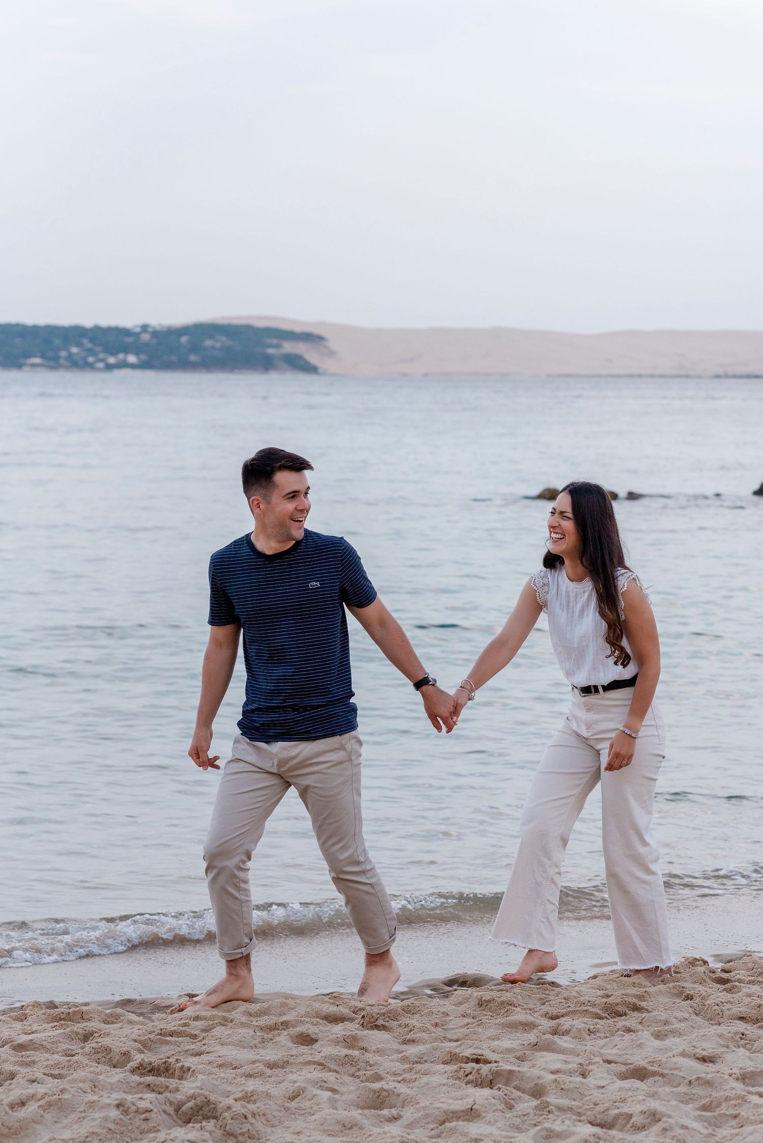 Playful couple walking and posing together on a sandy beach at sunset with ocean waves in the background.