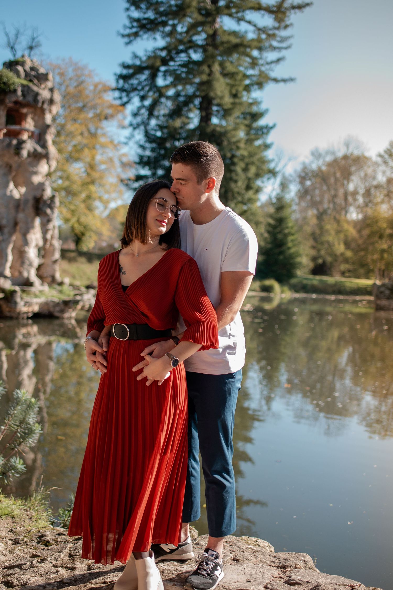Couple embracing by scenic pond with autumn trees and reflective water.
