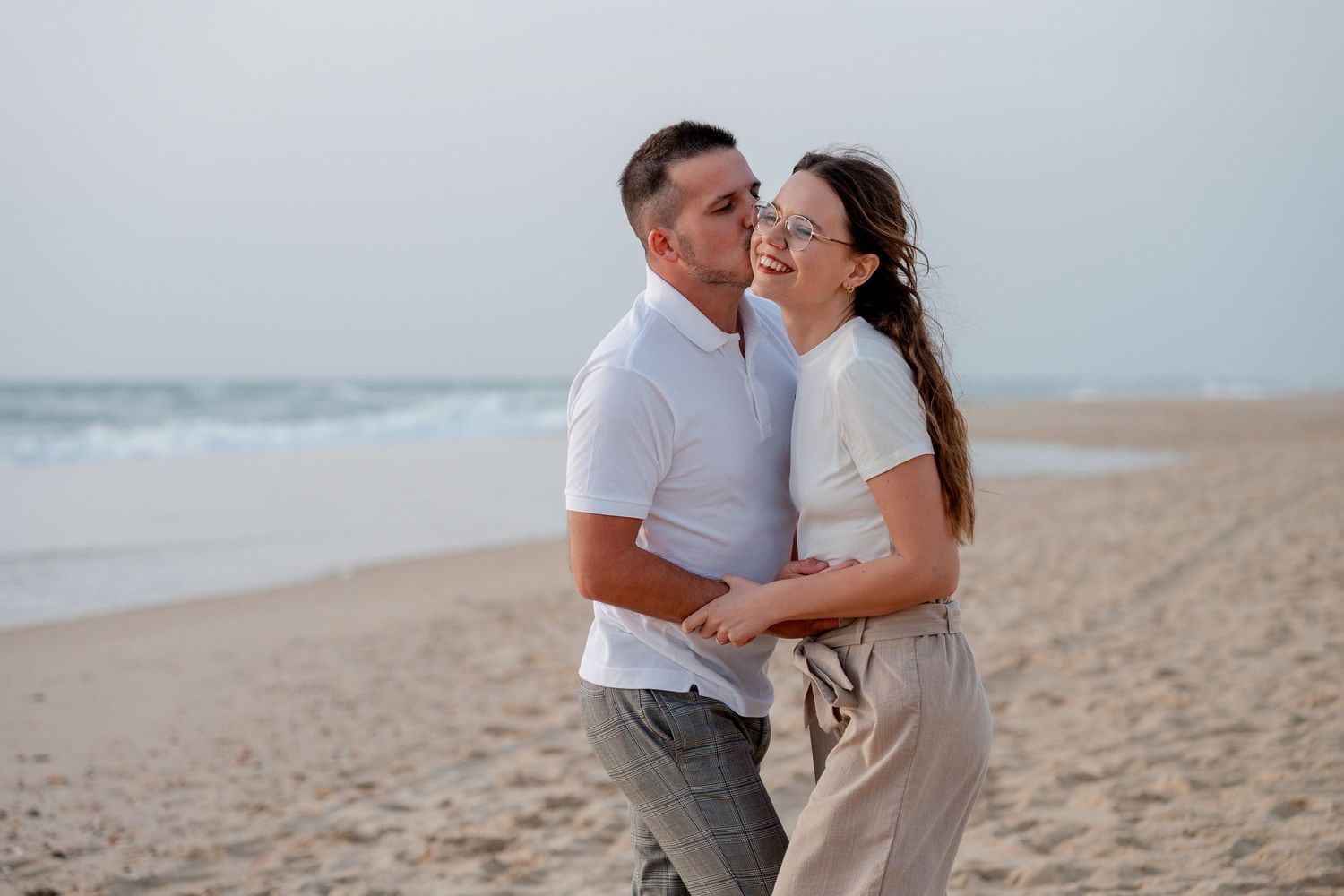 Romantic couple embracing and smiling on sandy beach with ocean waves in background.