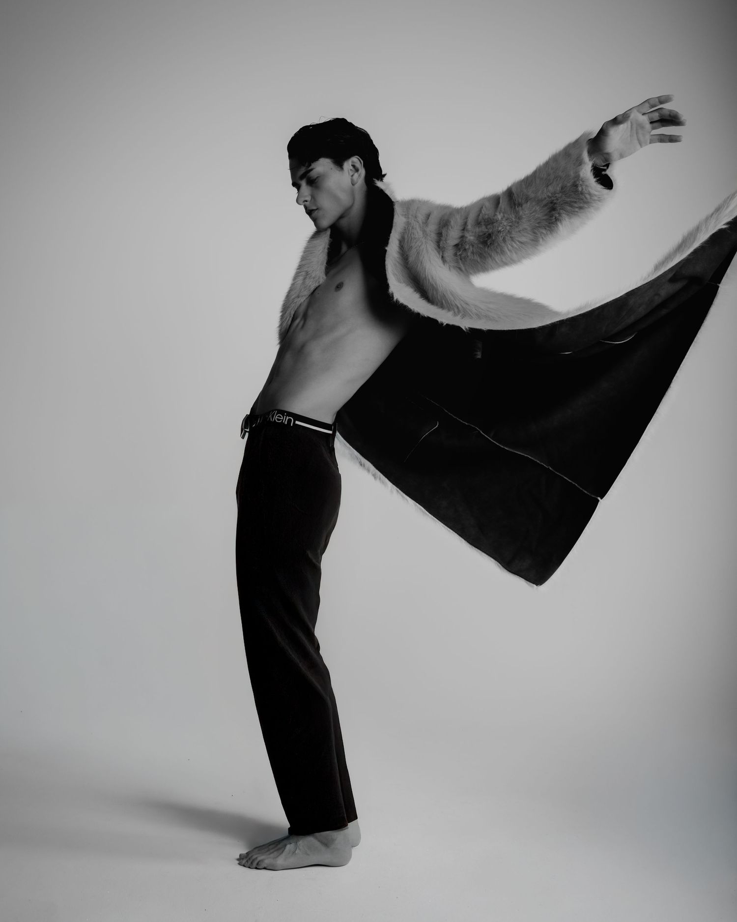 Shirtless male model poses dynamically in black trousers and fur coat in black and white editorial photo.