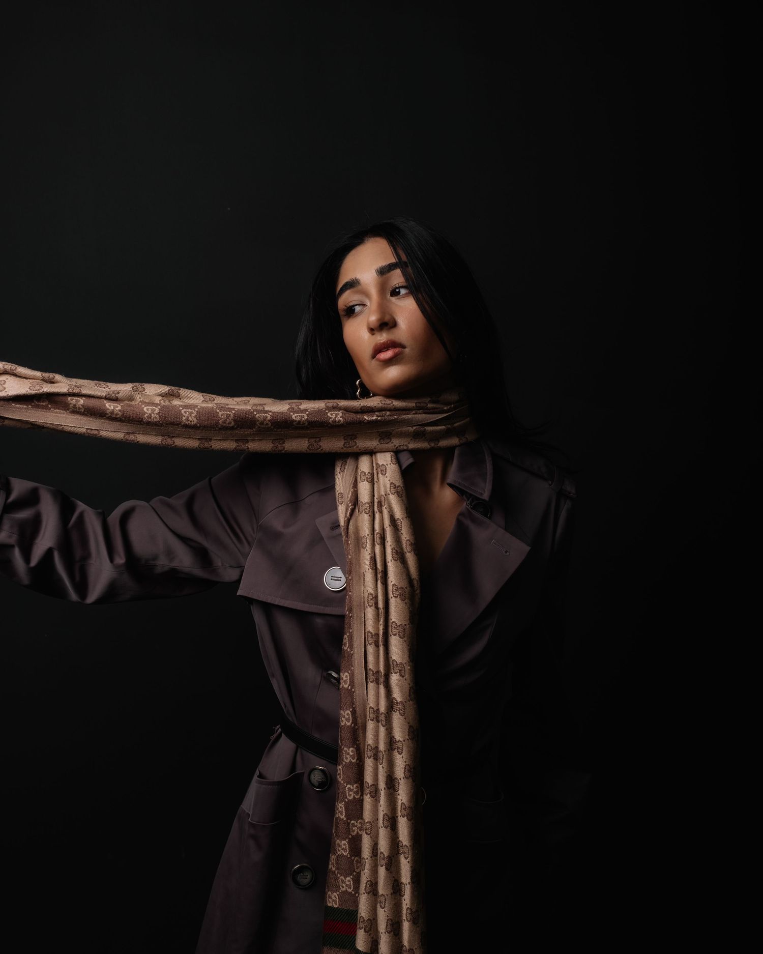 Dramatic portrait against a dark background with a long, patterned scarf and a brown trench coat in moody lighting.