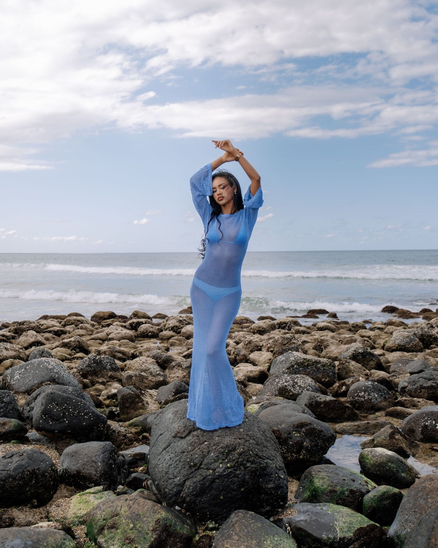 Woman in sheer blue knit maxi dress poses on rocky ocean shoreline with arms raised.