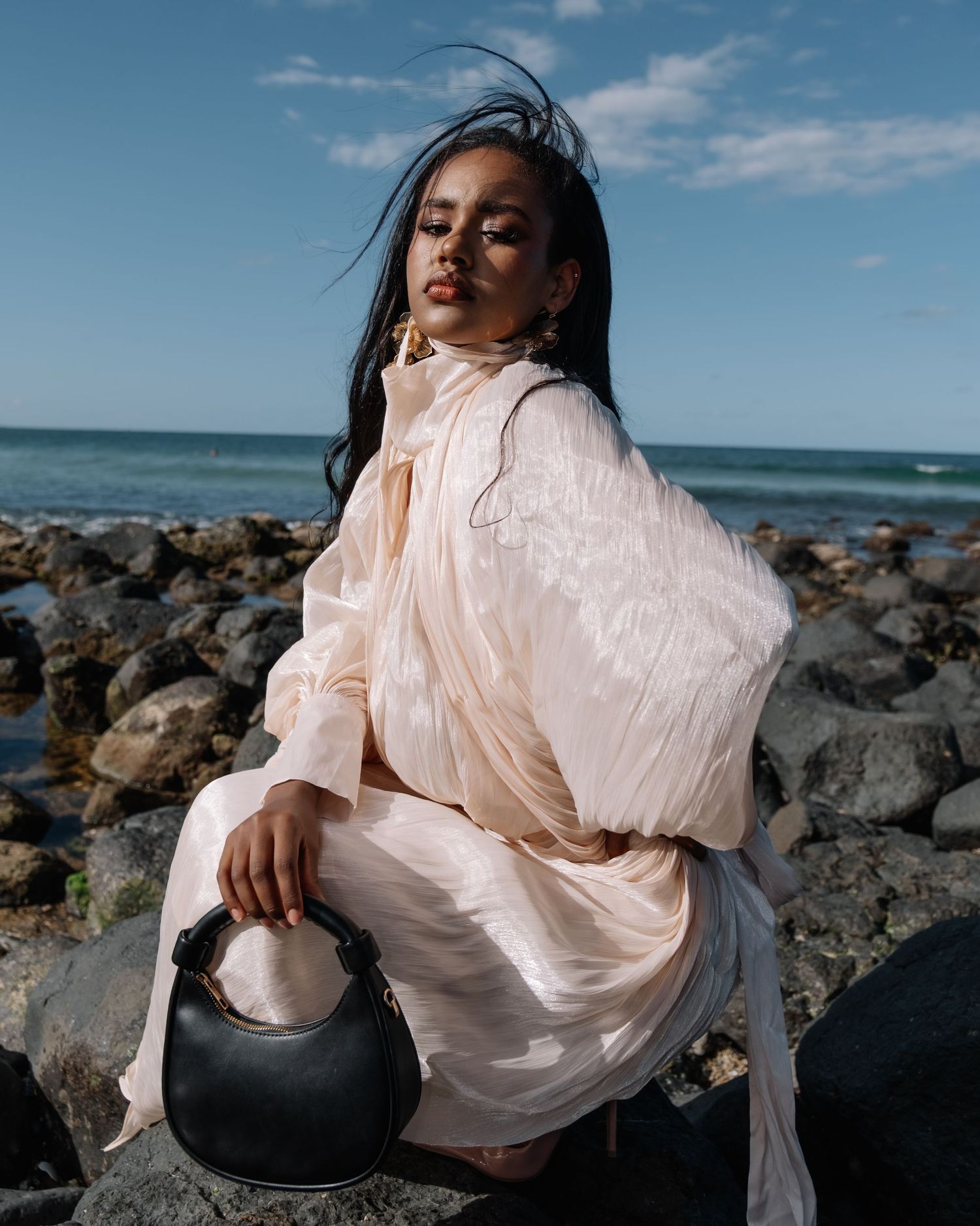 A figure in a flowing white dress poses gracefully against rocky coastal terrain, with the ocean and a blue sky in the background.