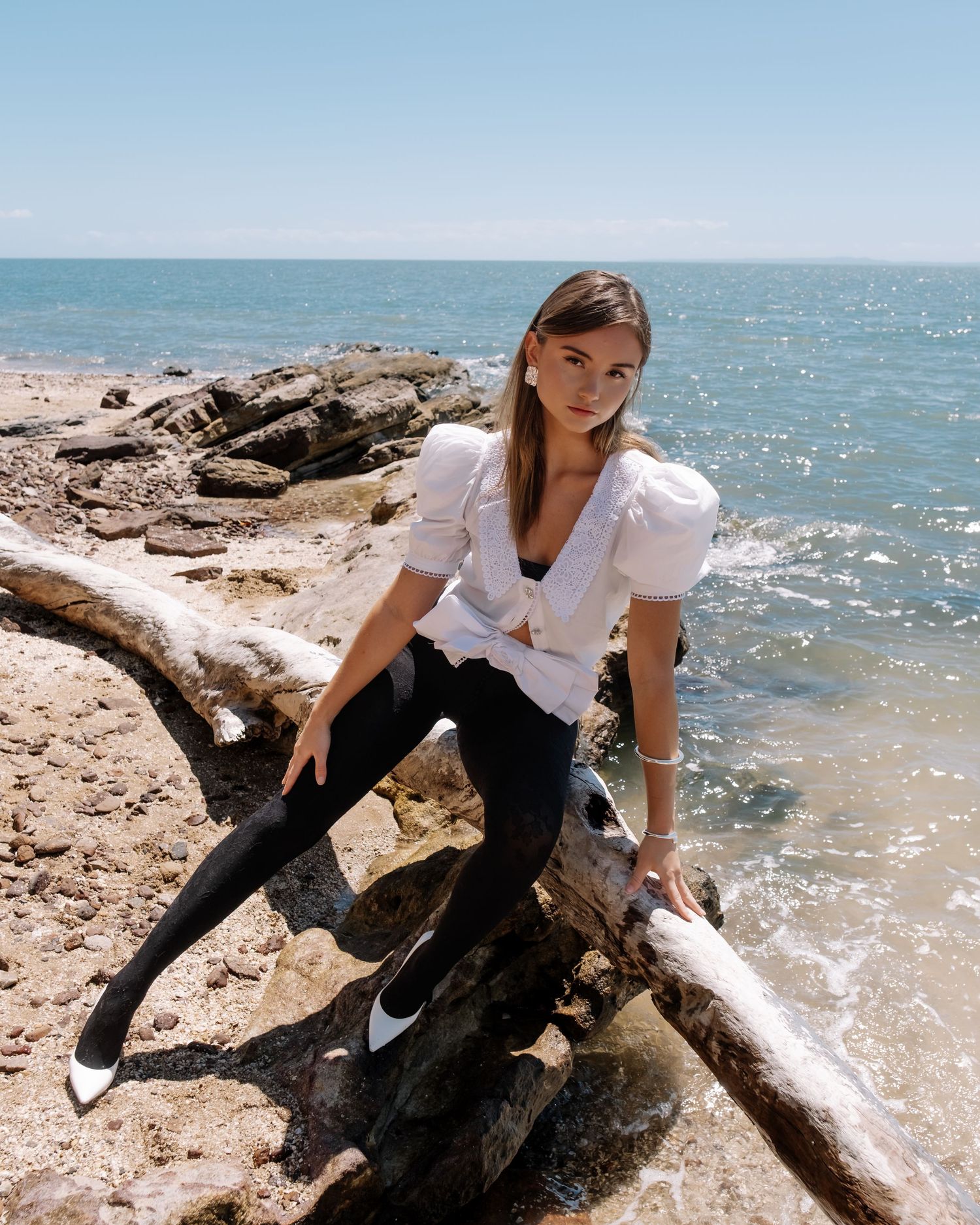 The subject, in casual attire, poses gracefully on rocky coastal terrain, with sparkling ocean waters in the background.