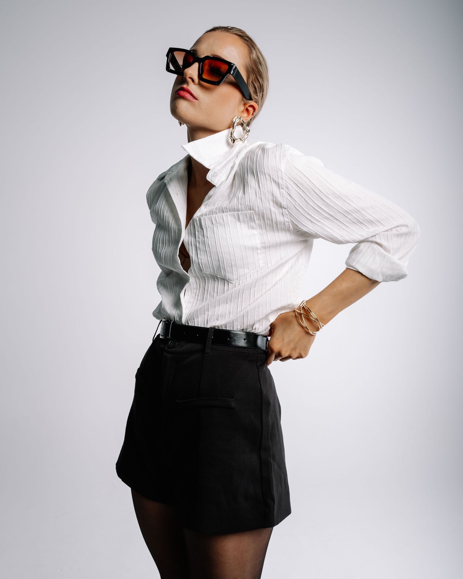 Blonde model wears white textured shirt, black mini skirt, tights and red sunglasses in studio shoot.