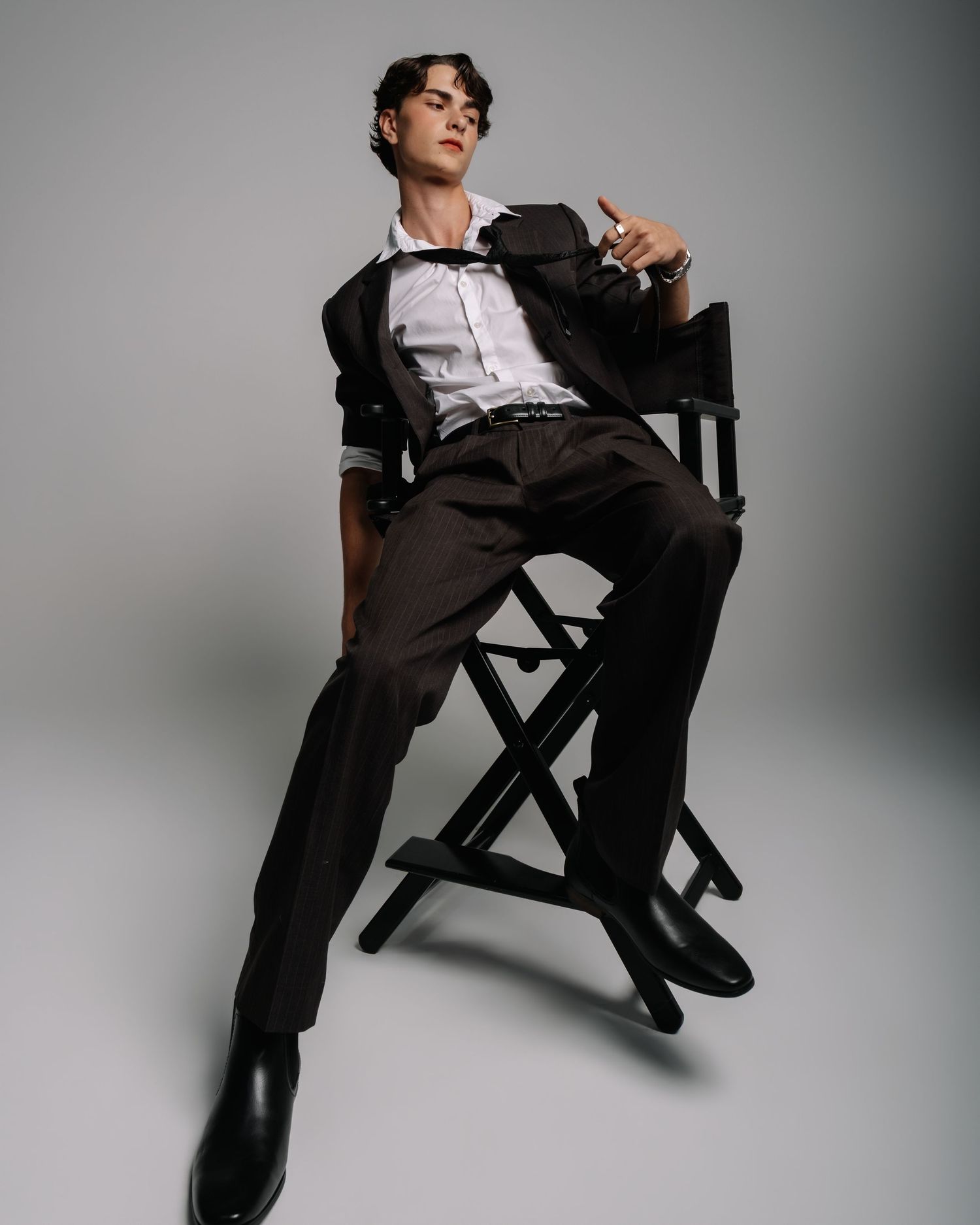 Male model sits in director's chair wearing dark suit with white shirt in moody studio portrait.