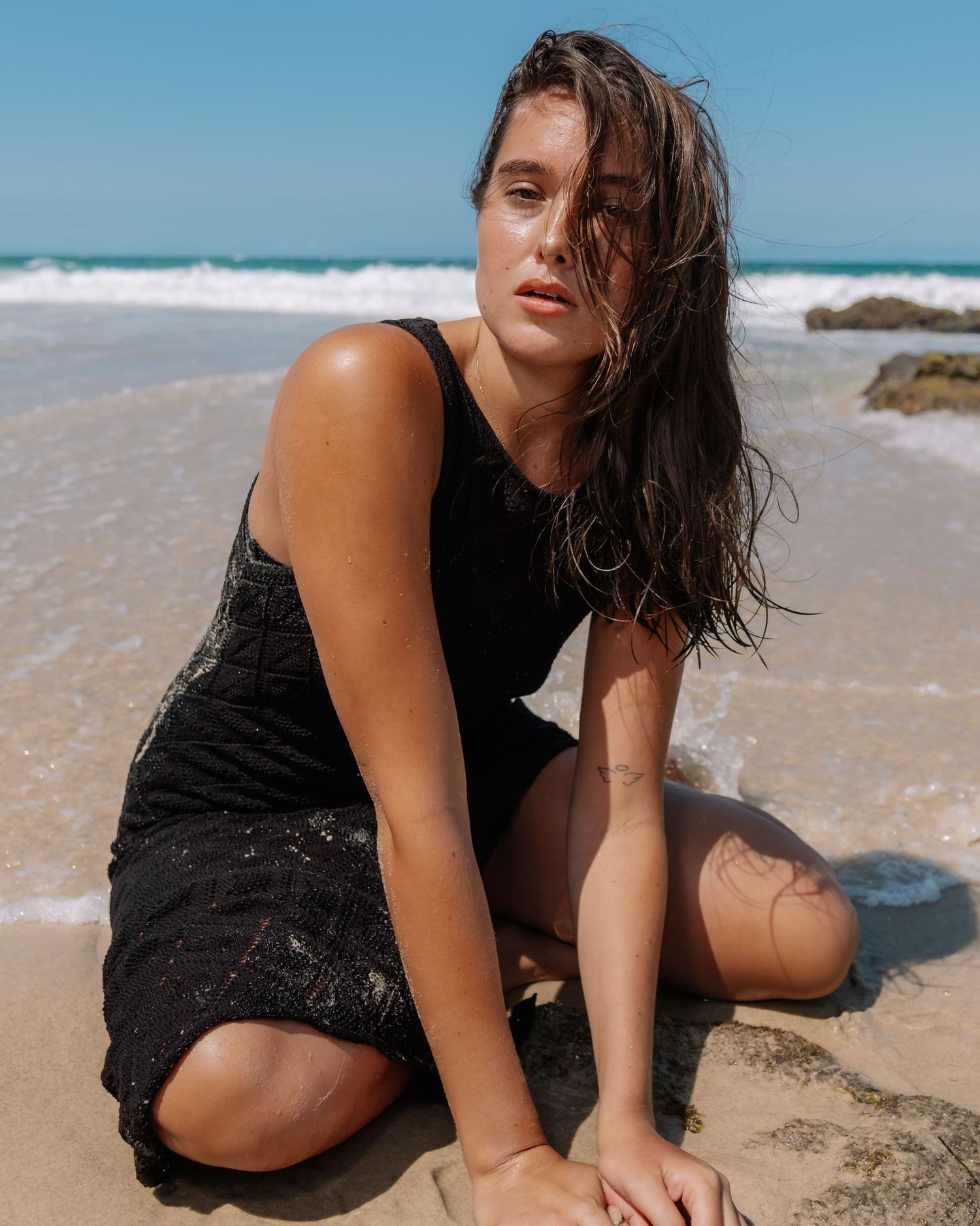 A person in a black dress poses on a sandy beach with ocean waves and rocks in the background.