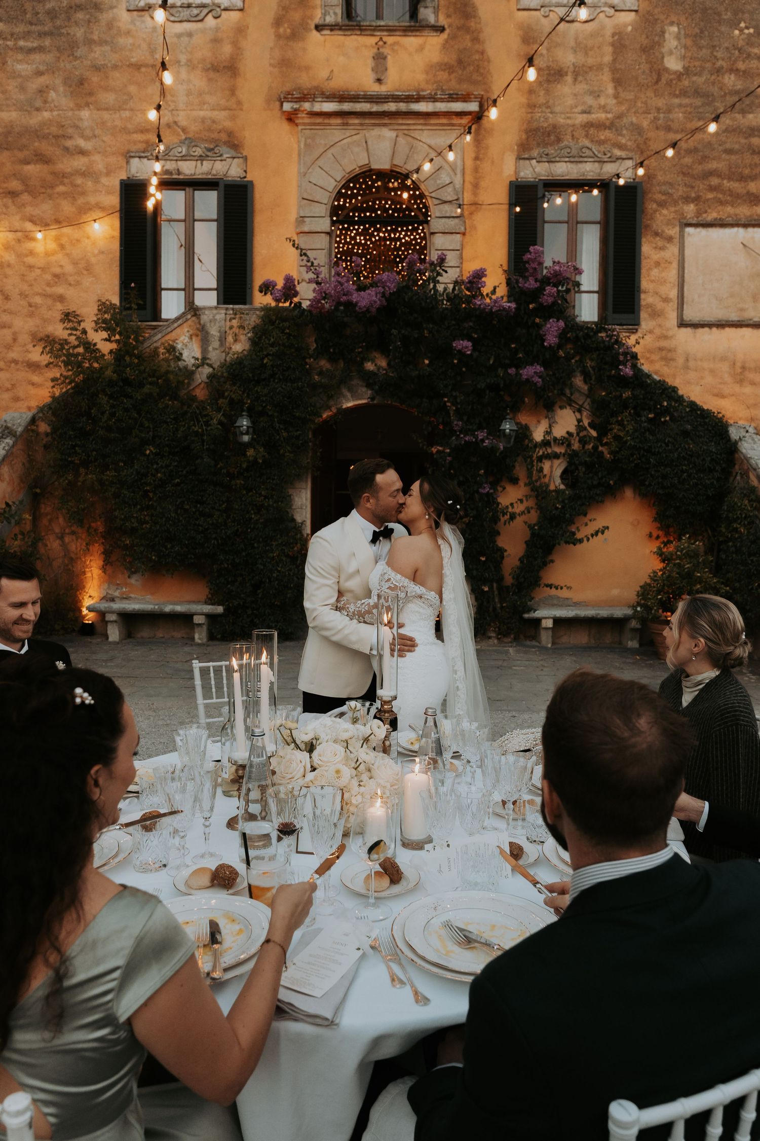 Wedding reception dinner tables set against rustic stone walls with string lights overhead.