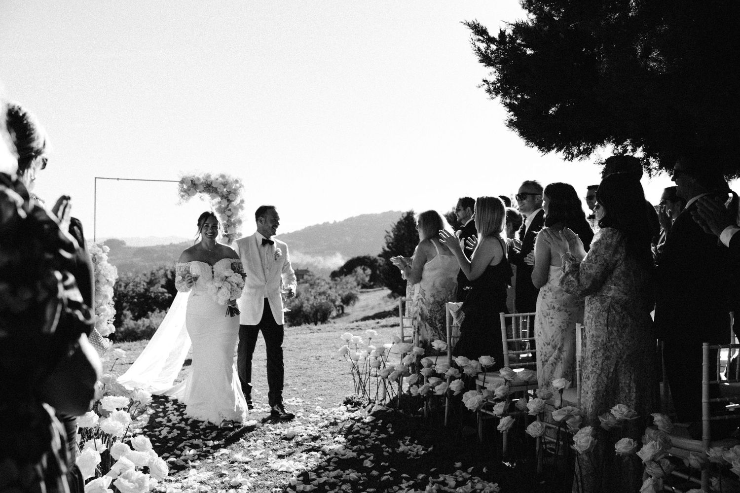 Black and white photo of a wedding ceremony with guests seated along a flower-lined pathway.