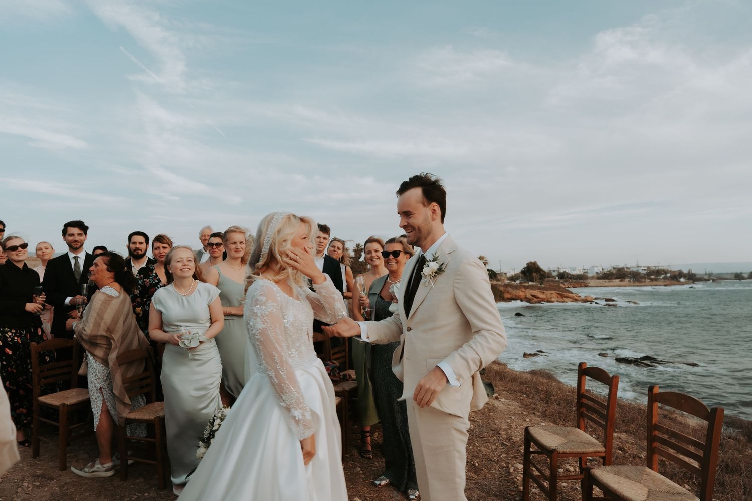 A seaside wedding ceremony with guests seated on wooden chairs overlooking the ocean.