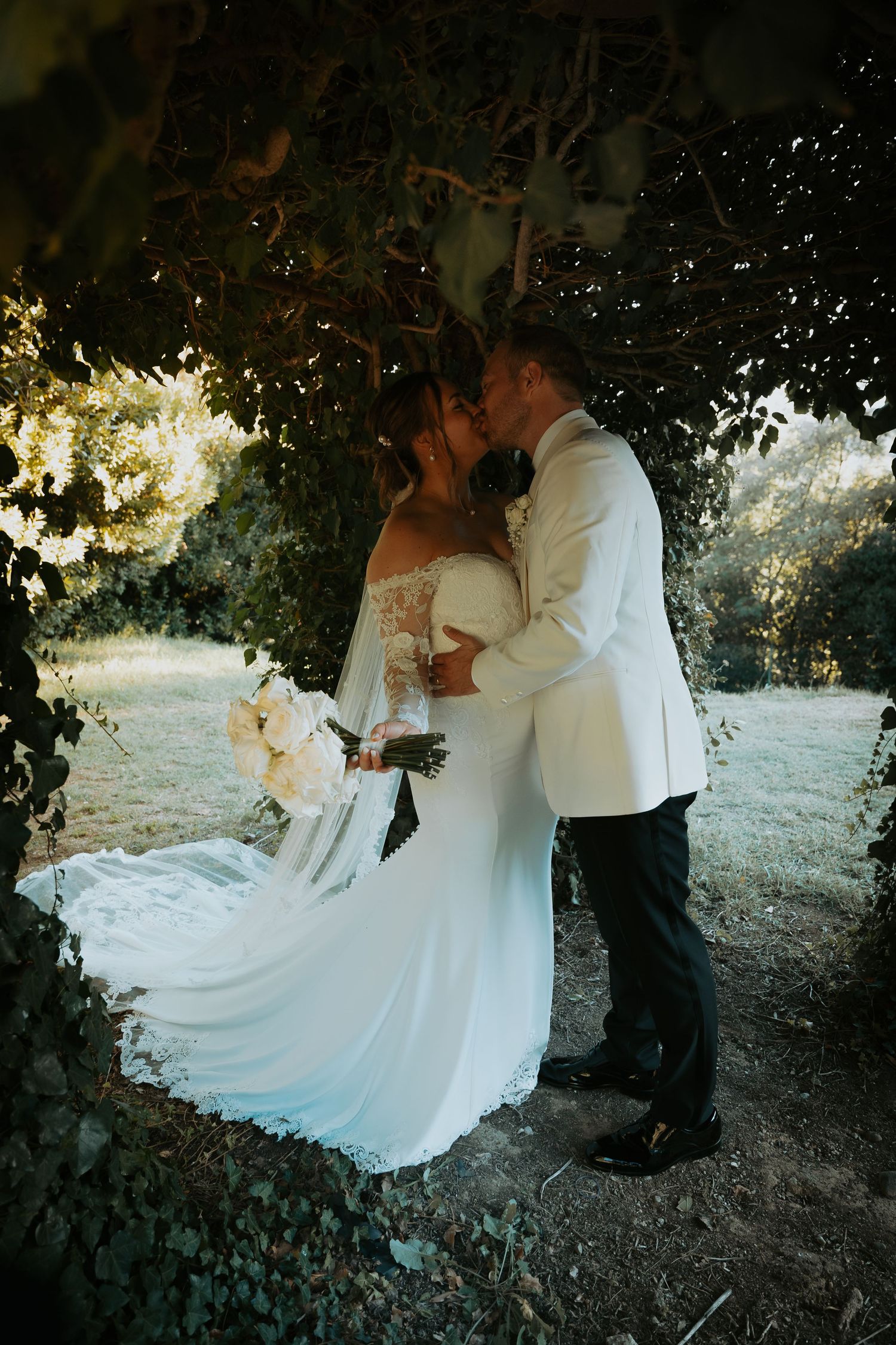 Newlyweds share an intimate kiss beneath tree branches at golden hour.