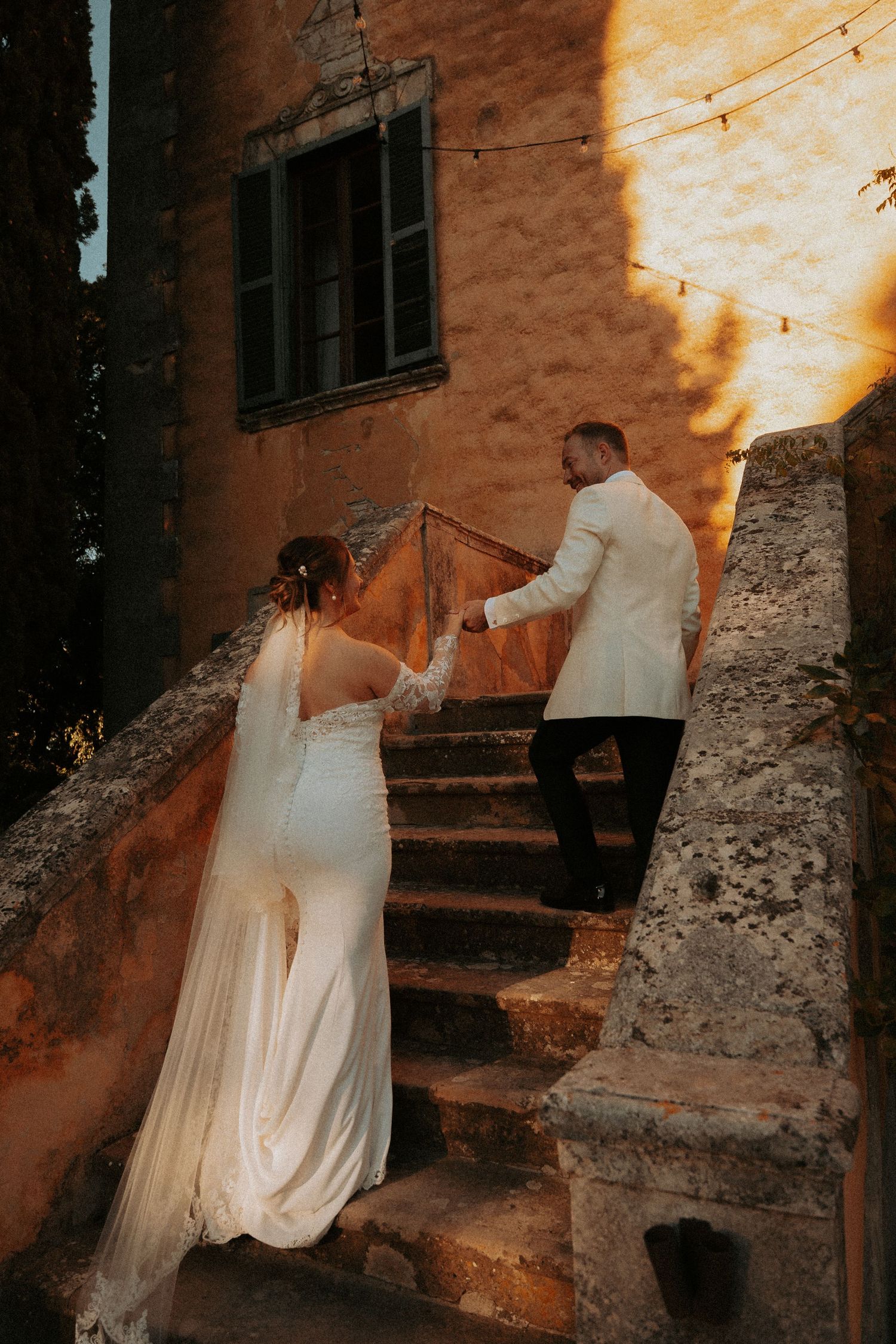 Couple in wedding attire climbing rustic stone steps at sunset.