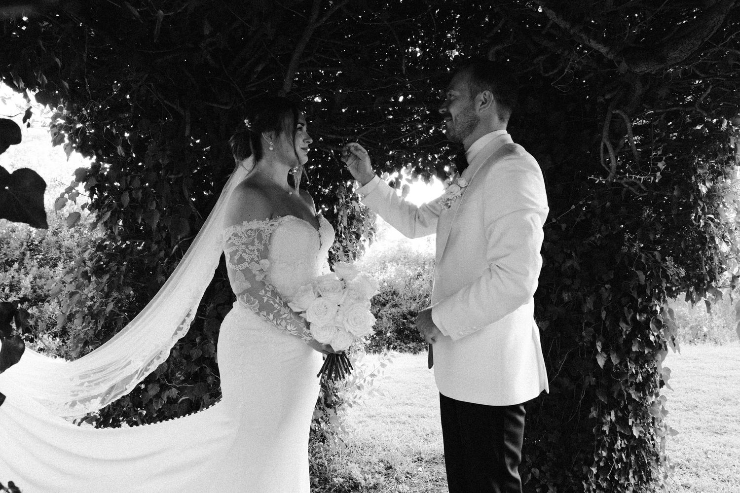 A romantic wedding moment captured in black and white under a natural tree canopy.