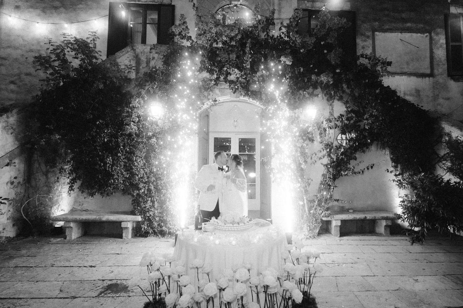 Black and white photo of an outdoor wedding venue illuminated by string lights at night.