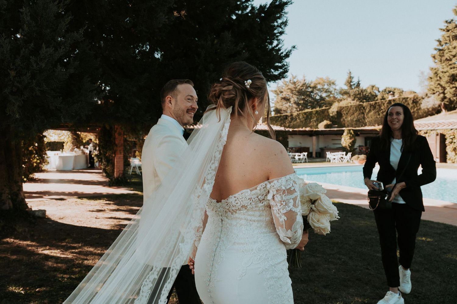Wedding couple walks past swimming pool area during outdoor celebration.