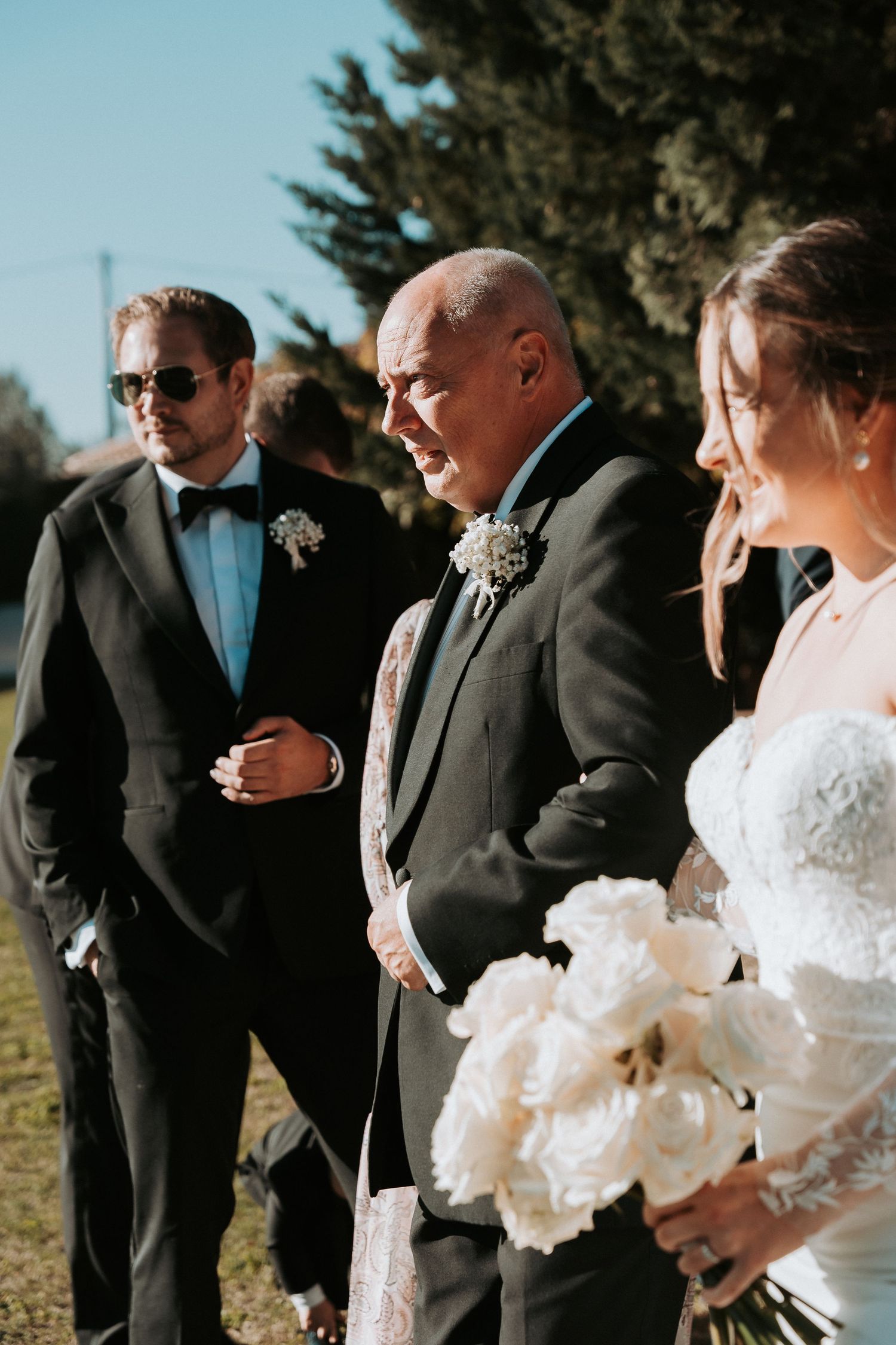 Wedding party members stand together outdoors wearing formal attire with a white bouquet.