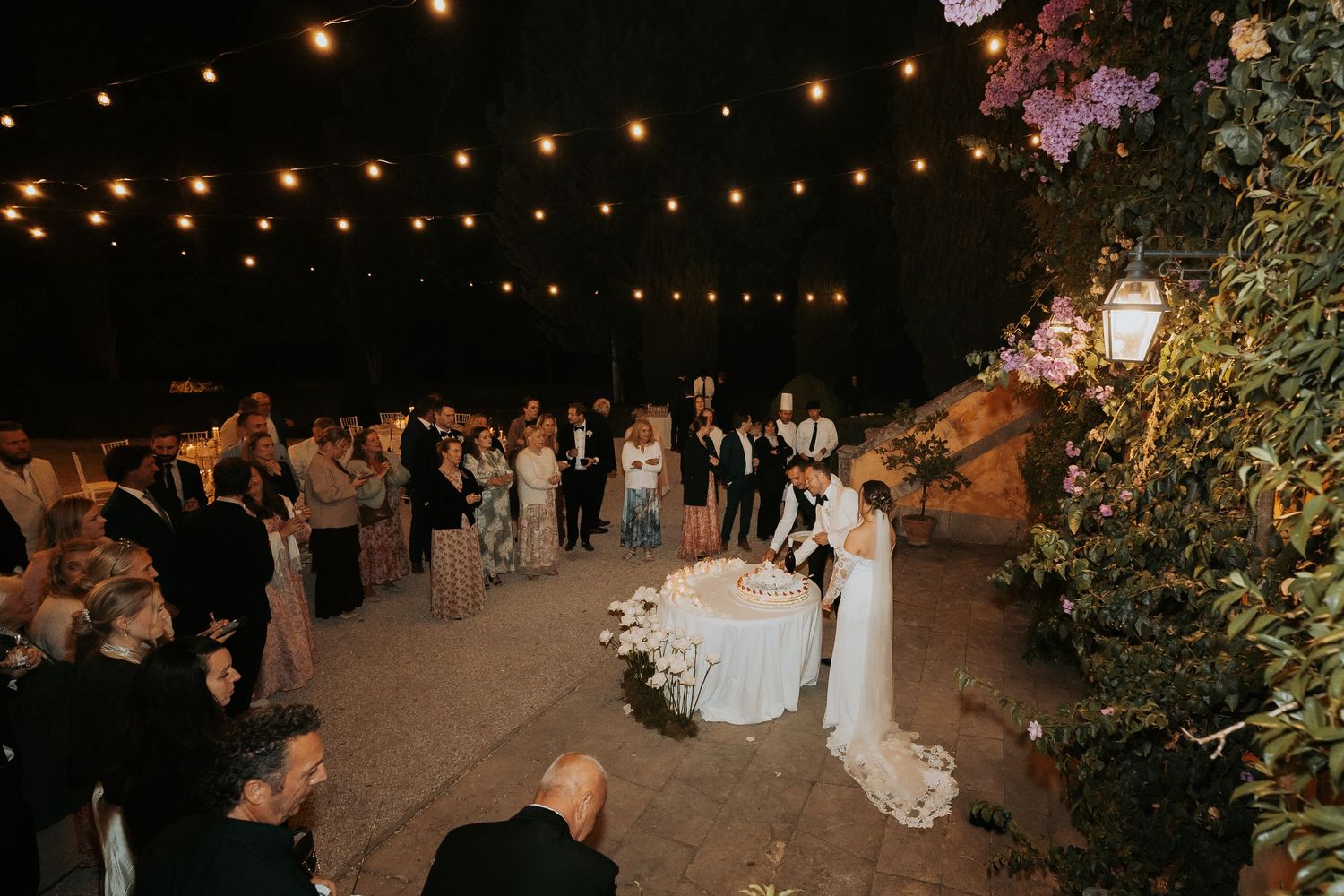 Wedding guests enjoying an evening celebration under twinkling string lights in an outdoor courtyard.