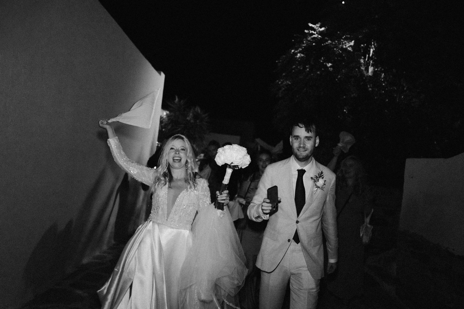 Newlyweds exit venue at night holding bouquets with dramatic lighting.