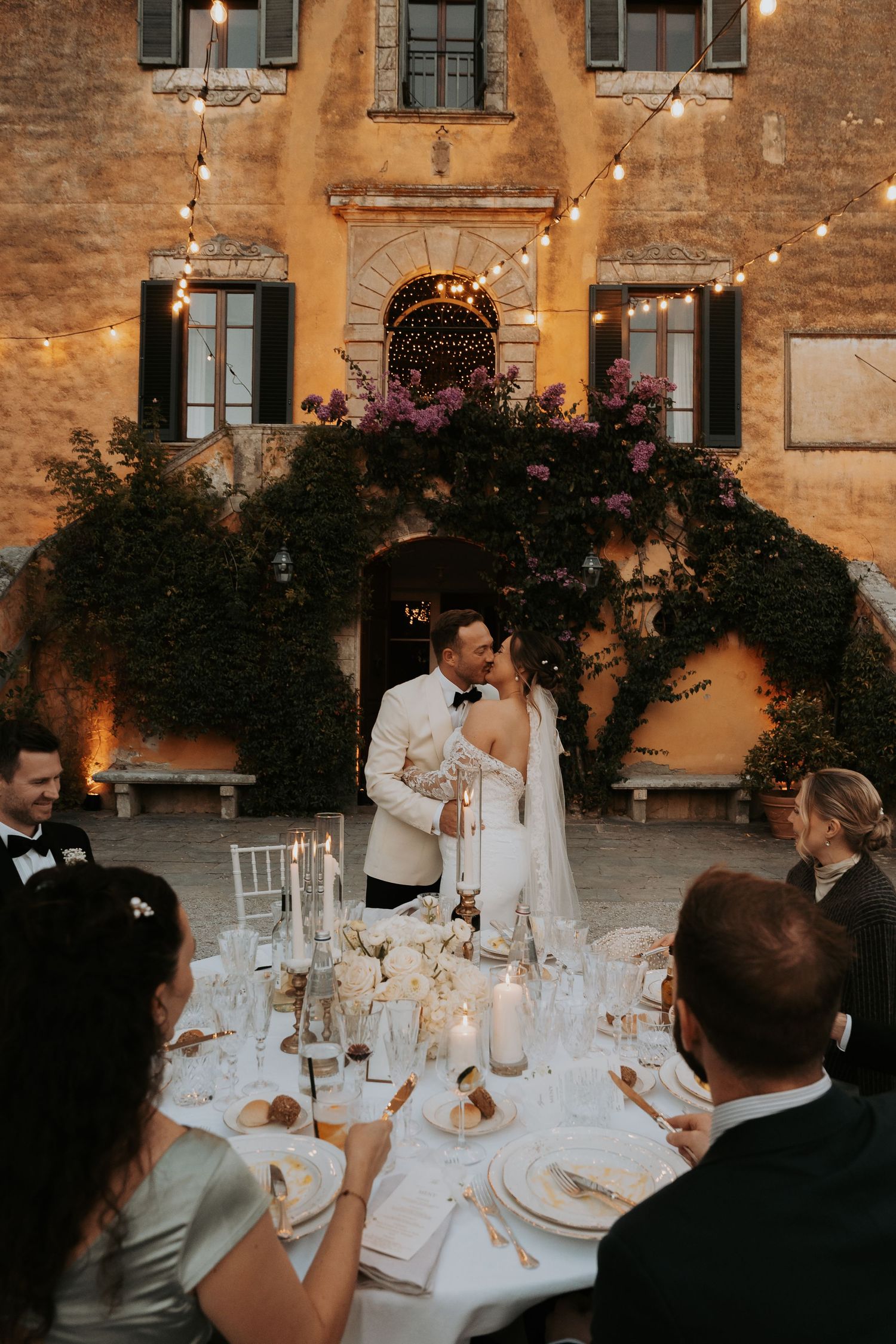 Wedding reception dinner at dusk with string lights and ivy-covered Italian villa backdrop.