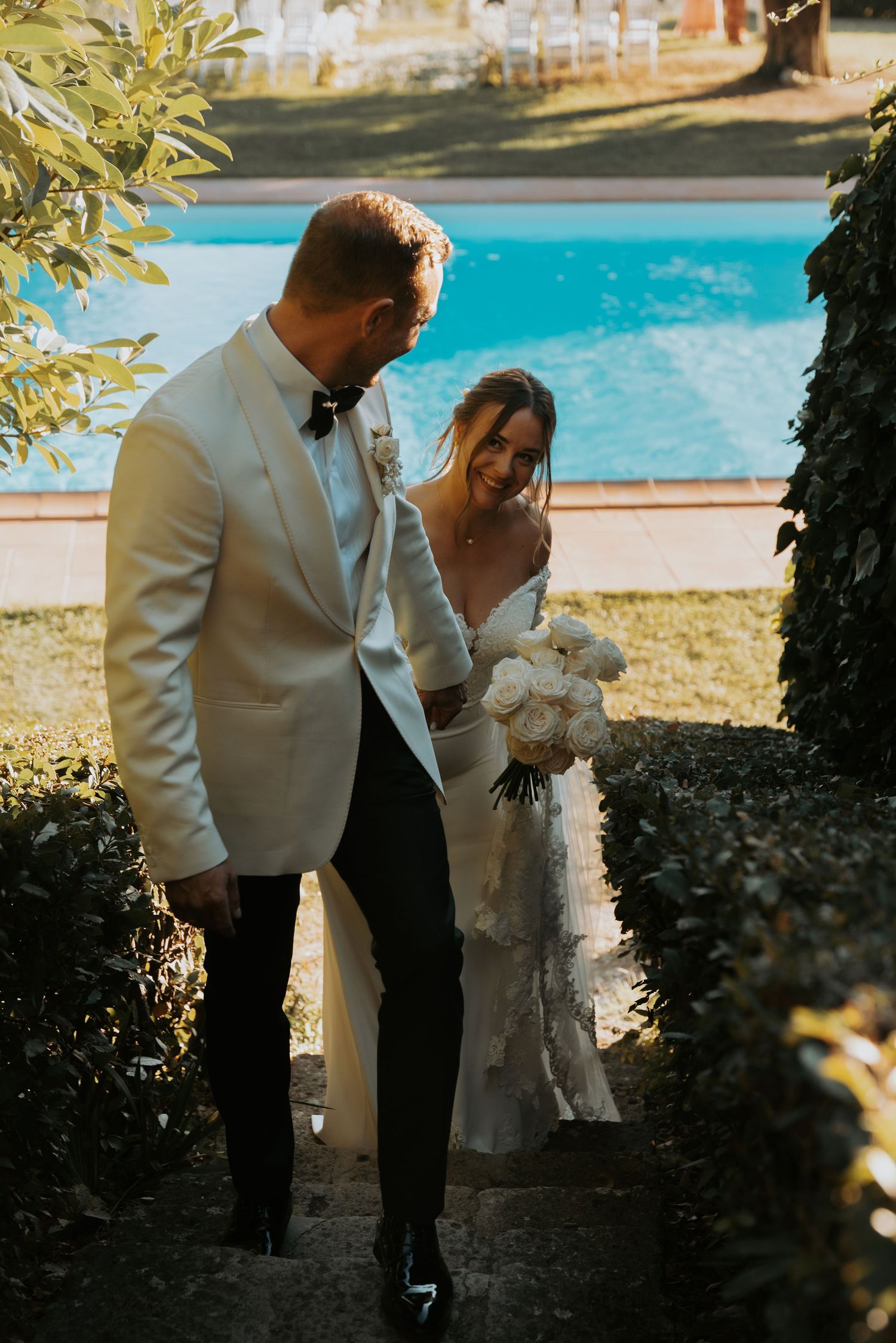 Elegant wedding couple descends stone steps near a swimming pool.