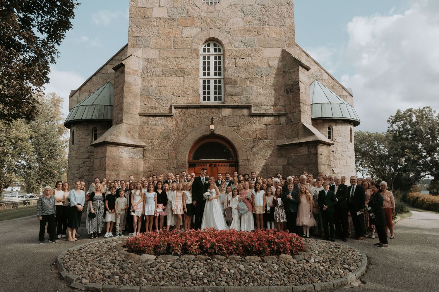 Large wedding party gathered outside stone church with arched entrance and flower bed in foreground.